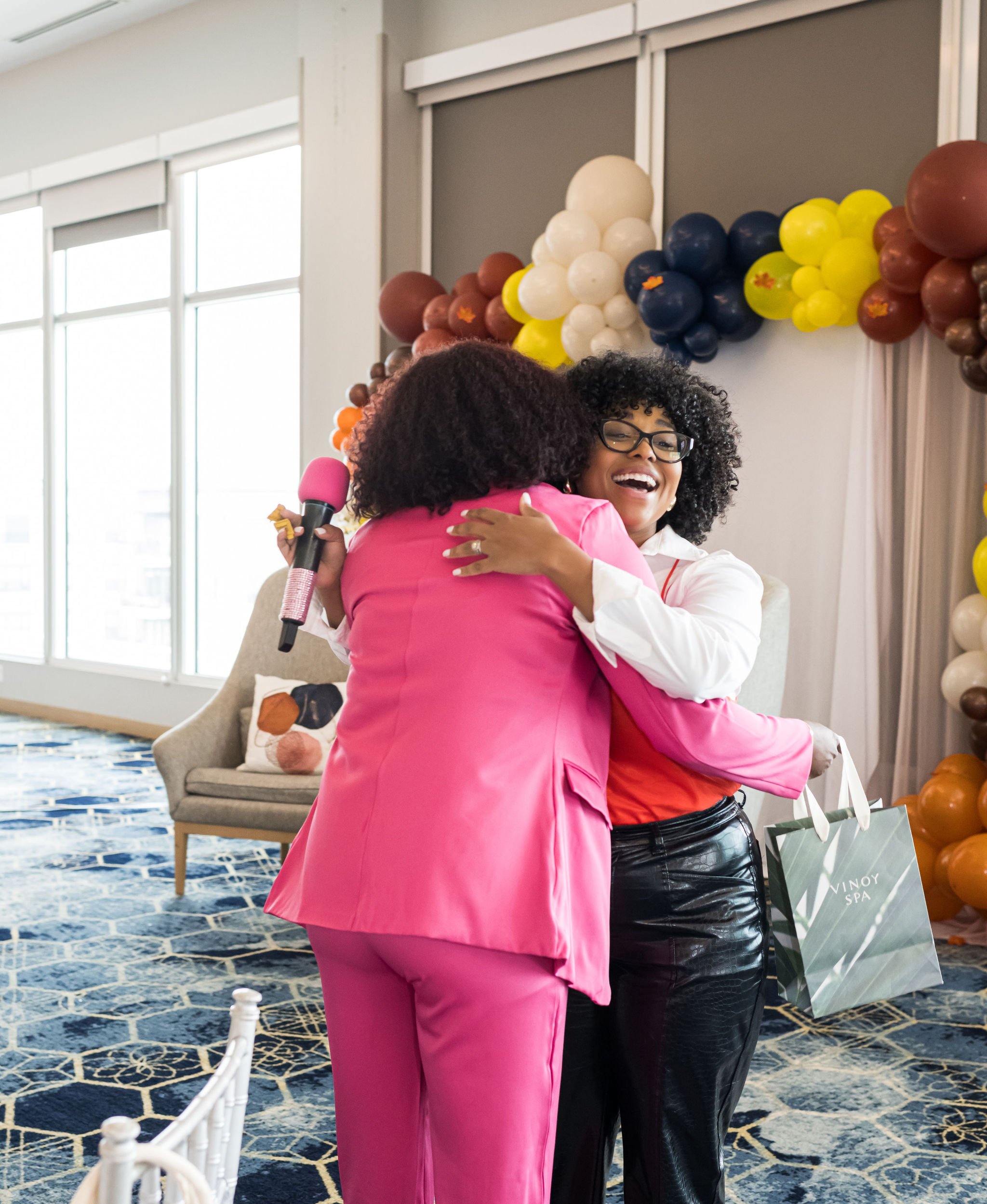 Two people hugging at an indoor event with colorful balloon decorations, one holding a microphone and the other holding a gift bag.