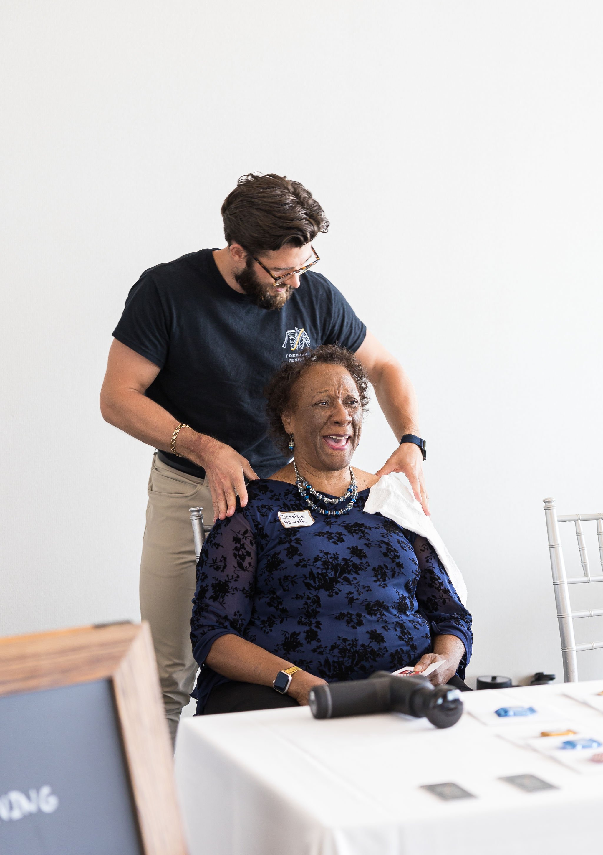 A man giving a chair massage to an older woman, who is smiling, in a well-lit room. The woman is seated at a table with small items on it, wearing a name tag and a blue floral top. The man is wearing glasses and a dark t-shirt.