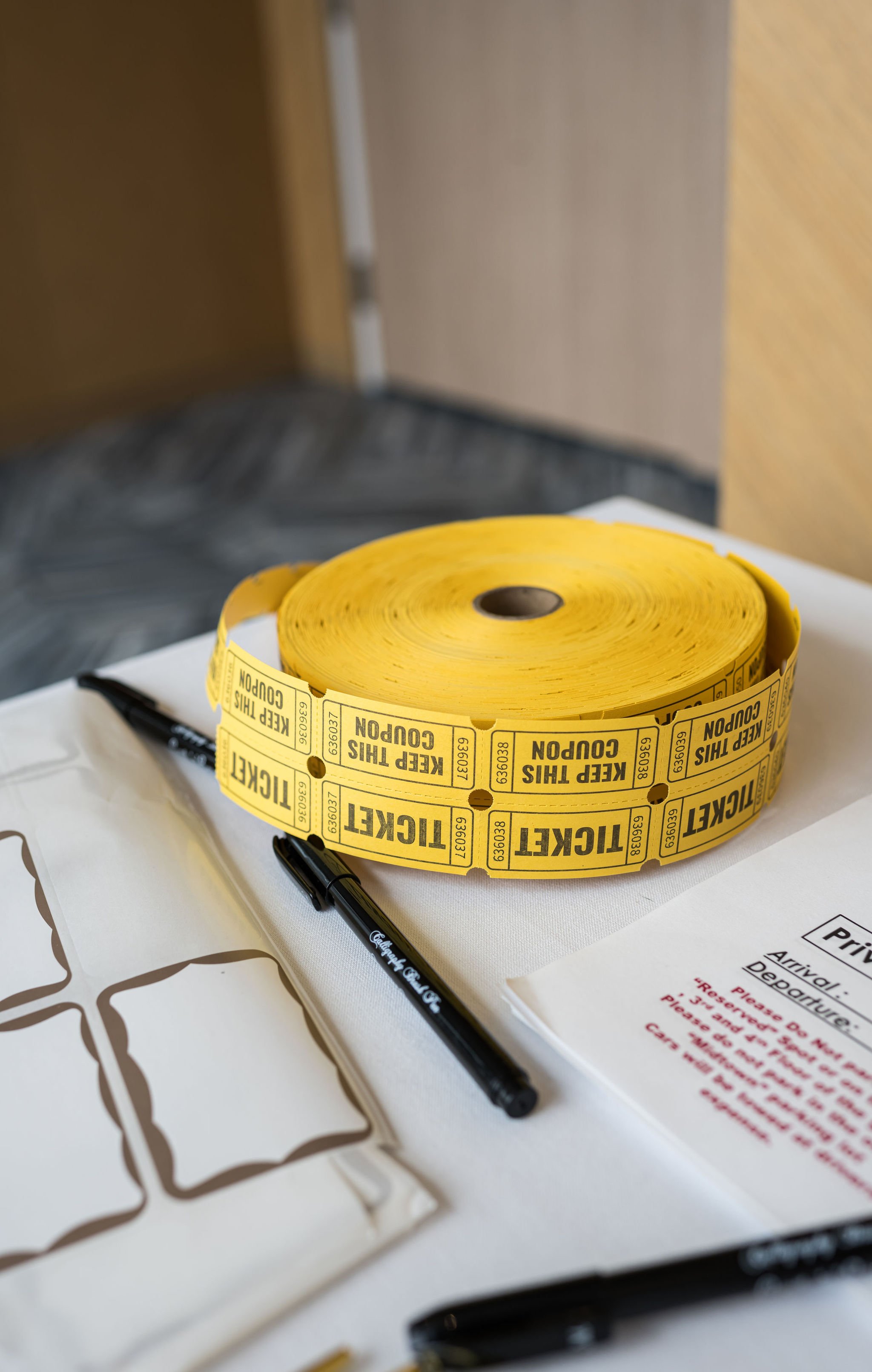 A roll of yellow "TICKET" raffle stubs with "KEEP THIS COUPON" printed on them, alongside black pens, blank labels, and printed instructions on a white surface.