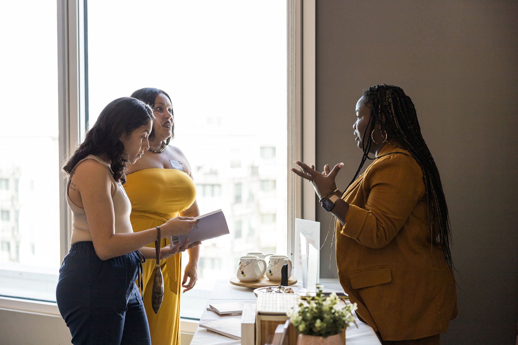 Three women engaging in a conversation near a window, one holding a book, with mugs and papers on a nearby table.