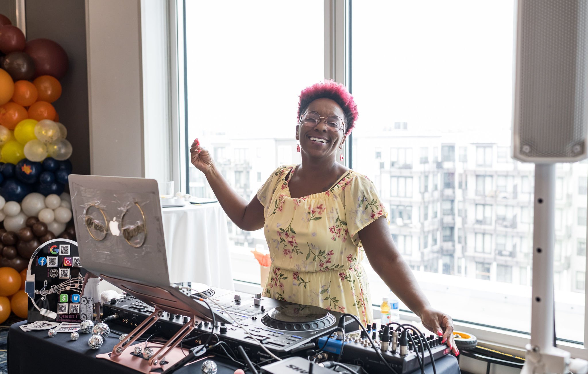 DJ smiling at a party with a laptop and mixing equipment on a table, balloons in the background, and a city view through the window.