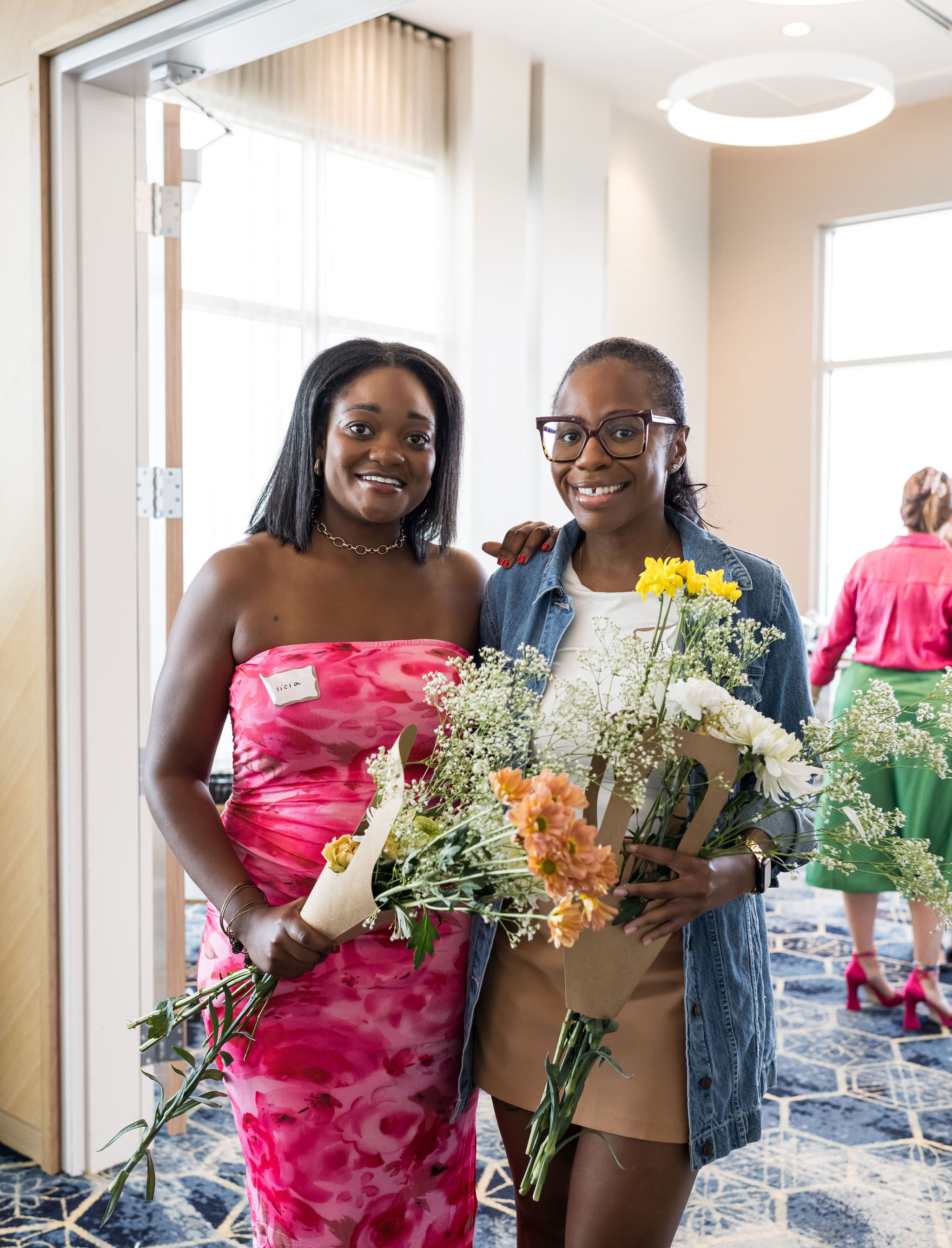 Two women smiling and holding bouquets of flowers, standing indoors with a modern, bright background. One wears a pink floral dress and the other in casual attire with glasses.