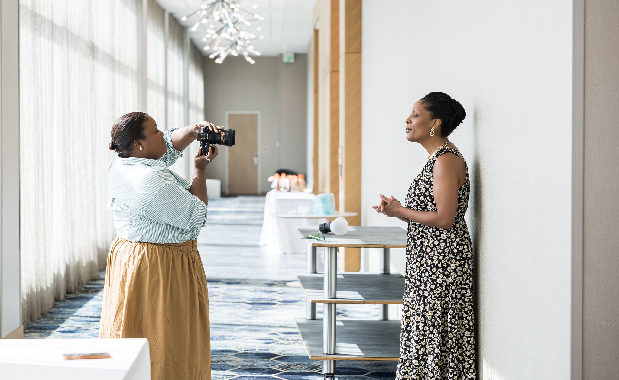 A photographer taking a picture of a woman leaning against a wall in a brightly lit corridor with modern decor.