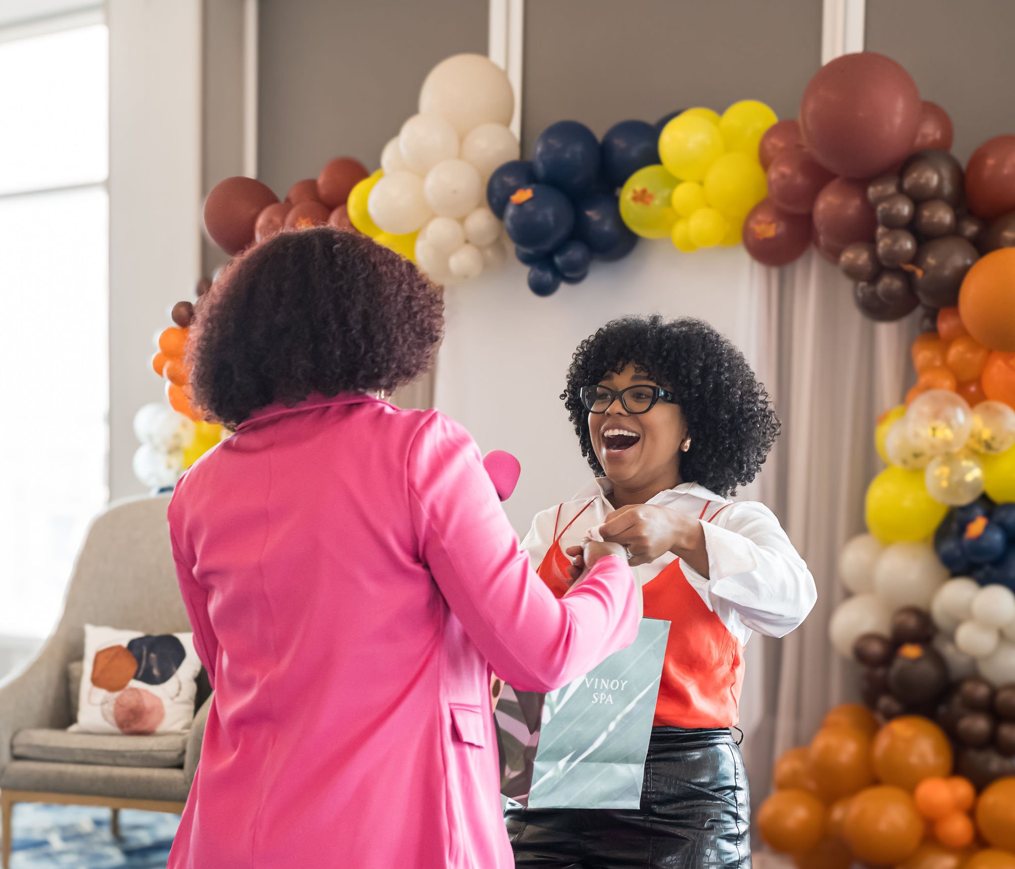 Two women exchanging a gift bag at an event with colorful balloon decorations in the background.