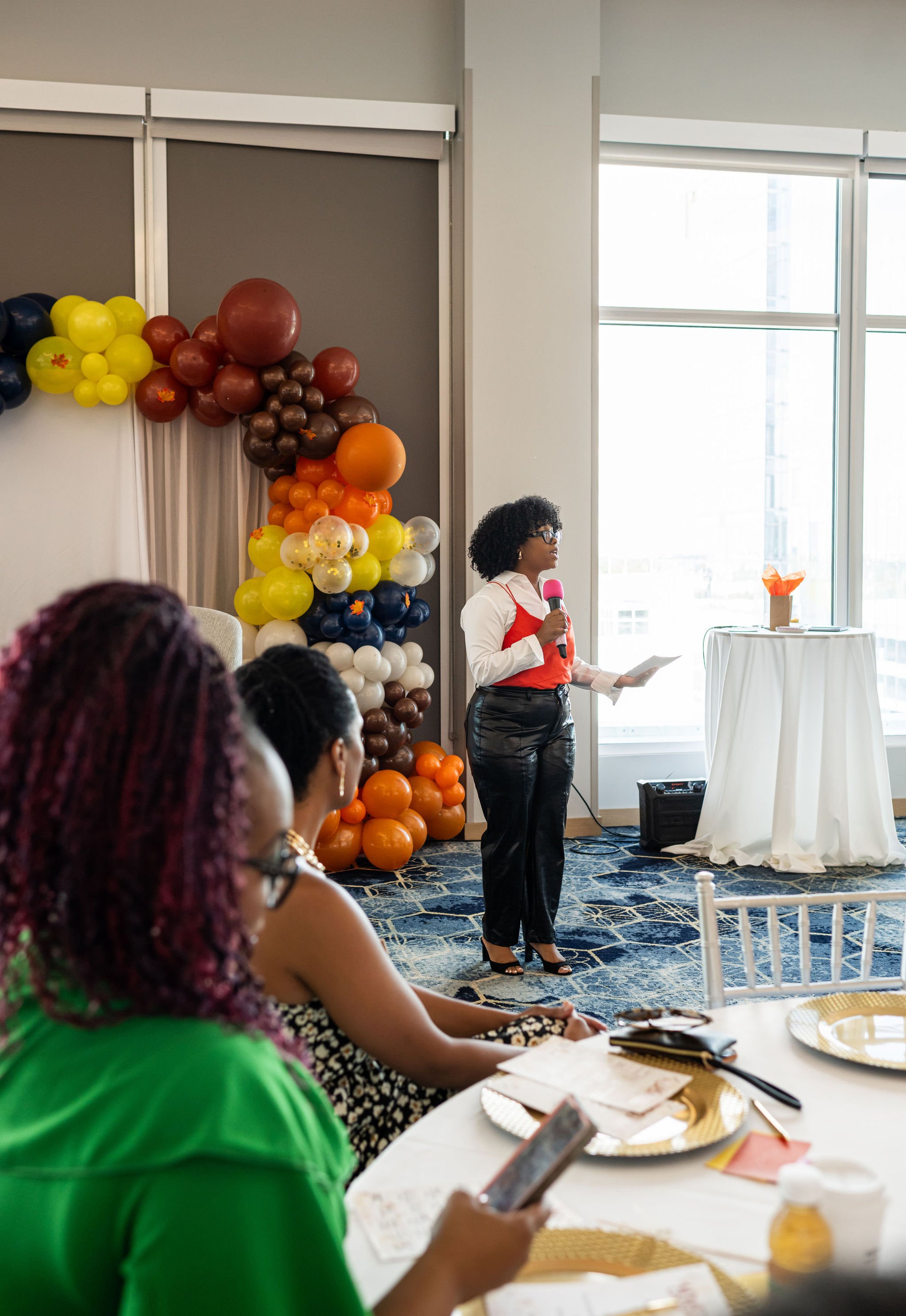 A woman speaking at an event with a colorful balloon arch in the background and attendees seated at a table in the foreground.