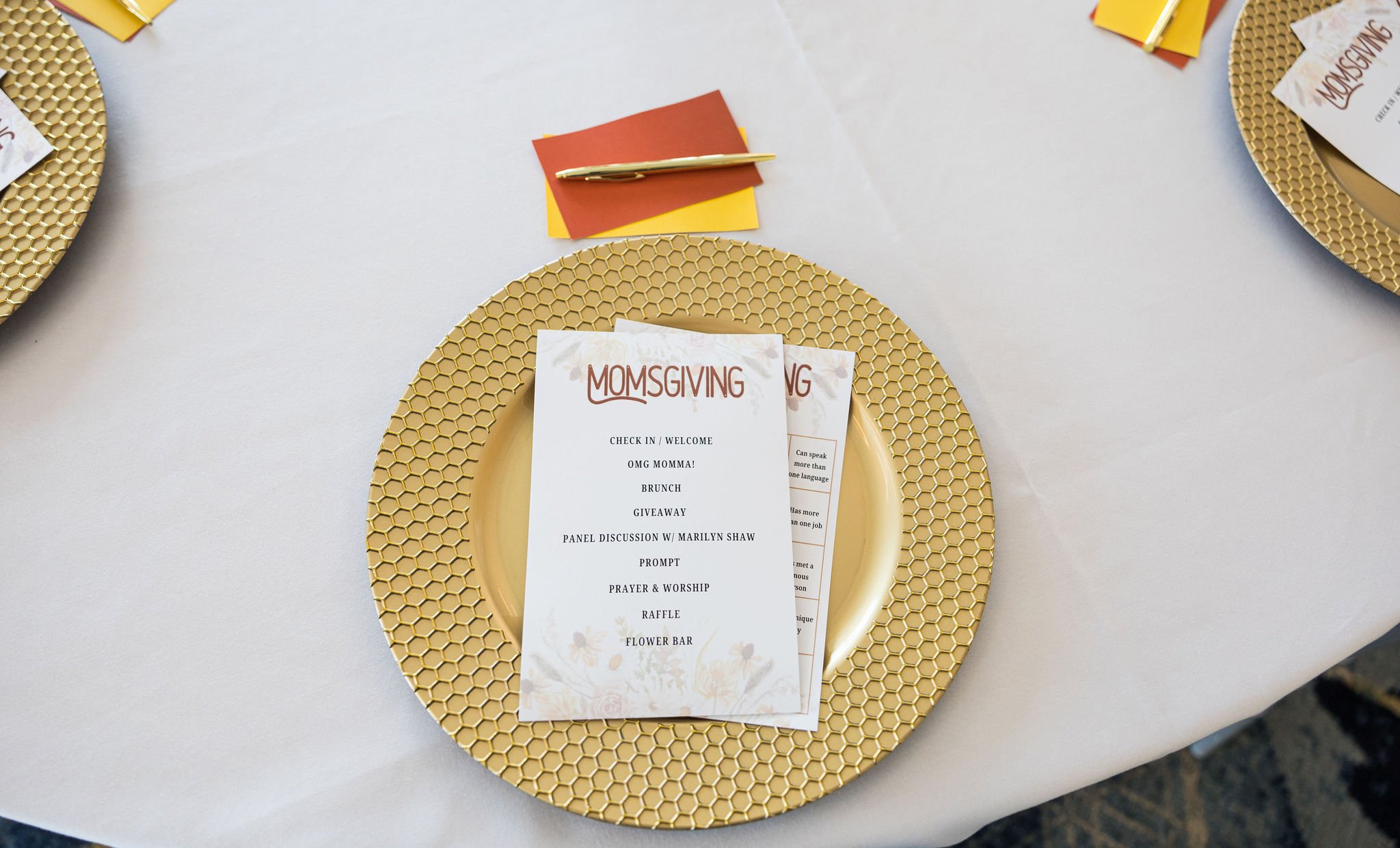 Elegant table setting with a honeycomb-patterned gold plate and a "MomsGiving" program on top. There are yellow and orange cards with a gold pen beside it on a white tablecloth.