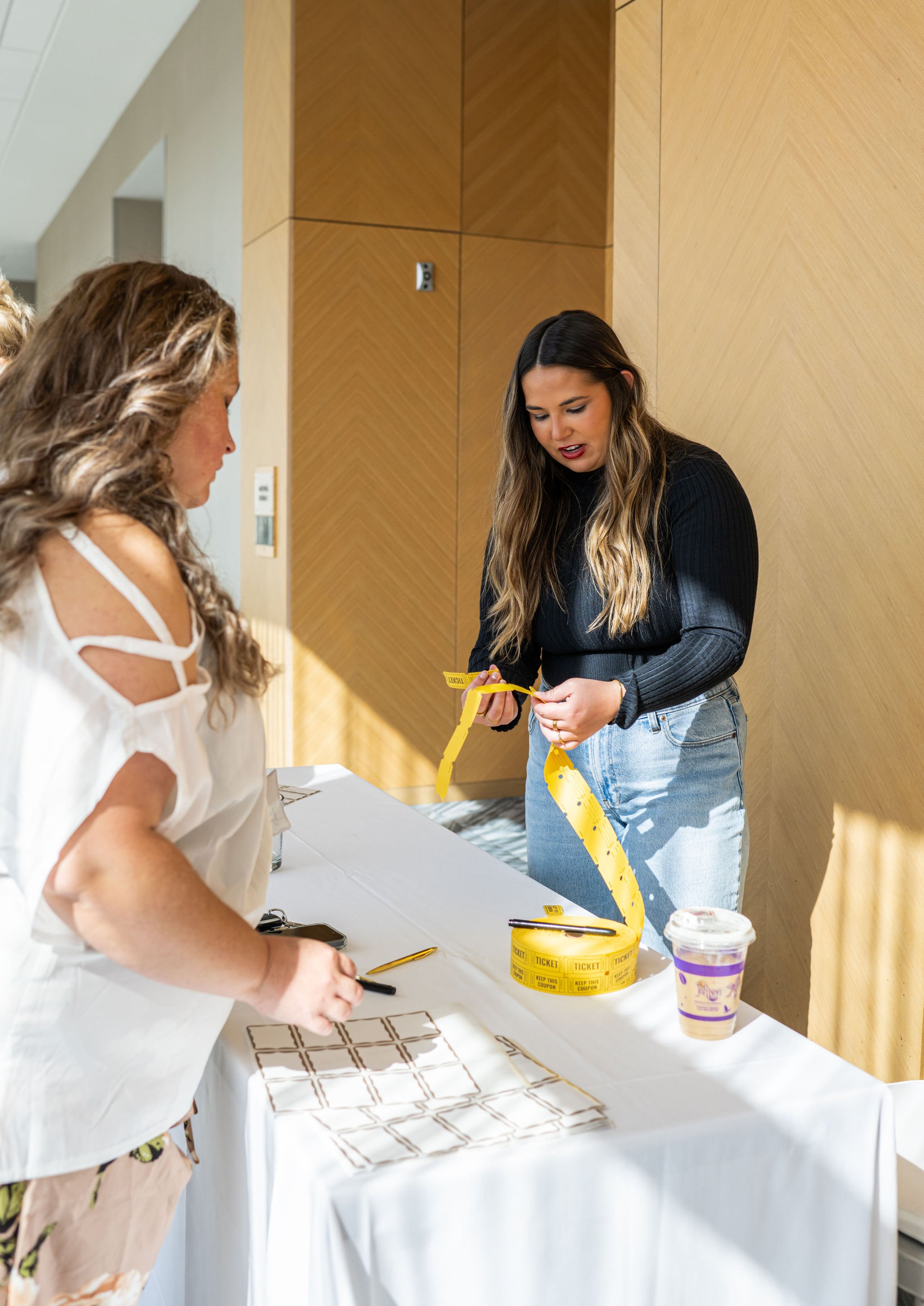 Two women at a table with yellow tickets and a drink in a cup.
