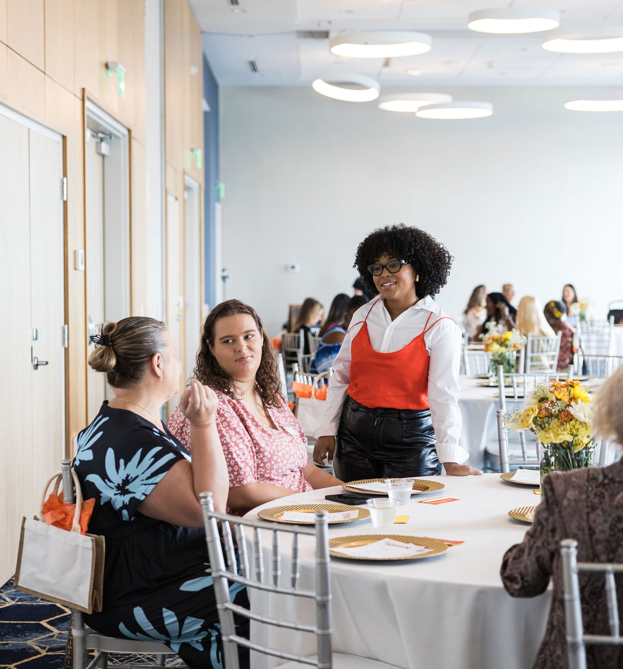 A woman in a red top and glasses stands next to a table having a conversation with two seated women. The room is set up for a formal event with round tables, white tablecloths, plates, and floral centerpieces. More people are seated at other tables i