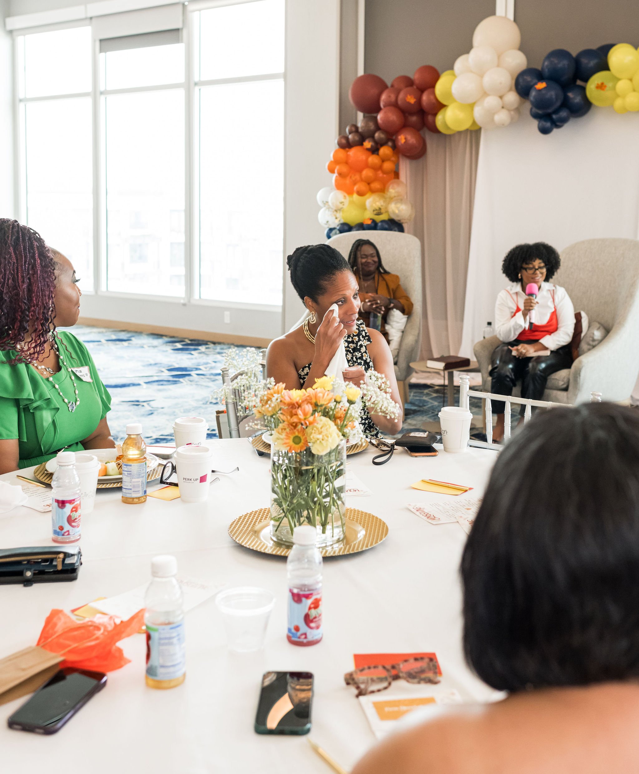 A diverse group of people sitting around a table engaged in discussion. The table is set with drinks, flower arrangements, and small plates. There is a balloon decoration in the background and a person holding a microphone, suggesting a casual event