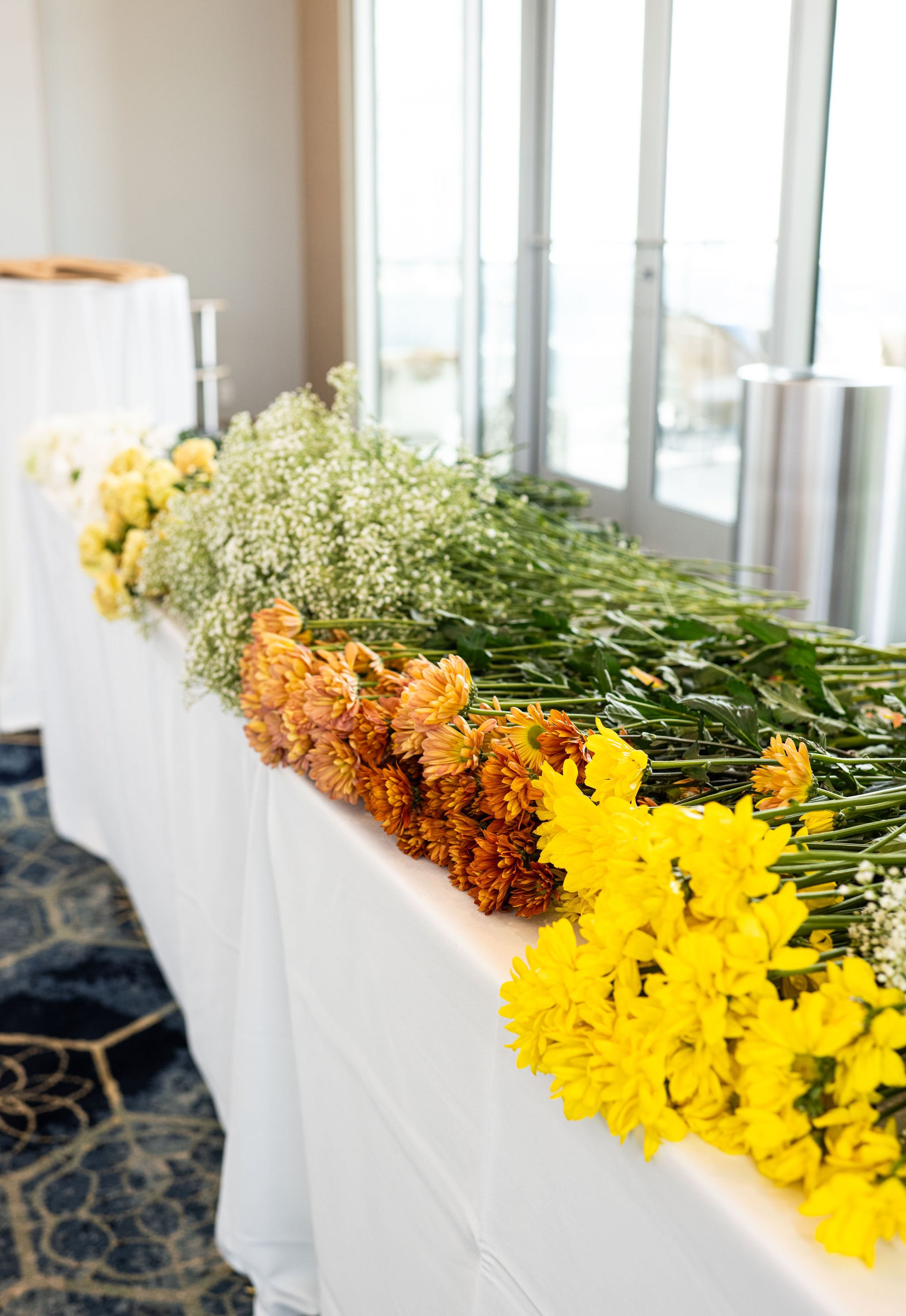 Assorted flowers, including yellow and orange blooms, laid on a table with a white tablecloth indoors.