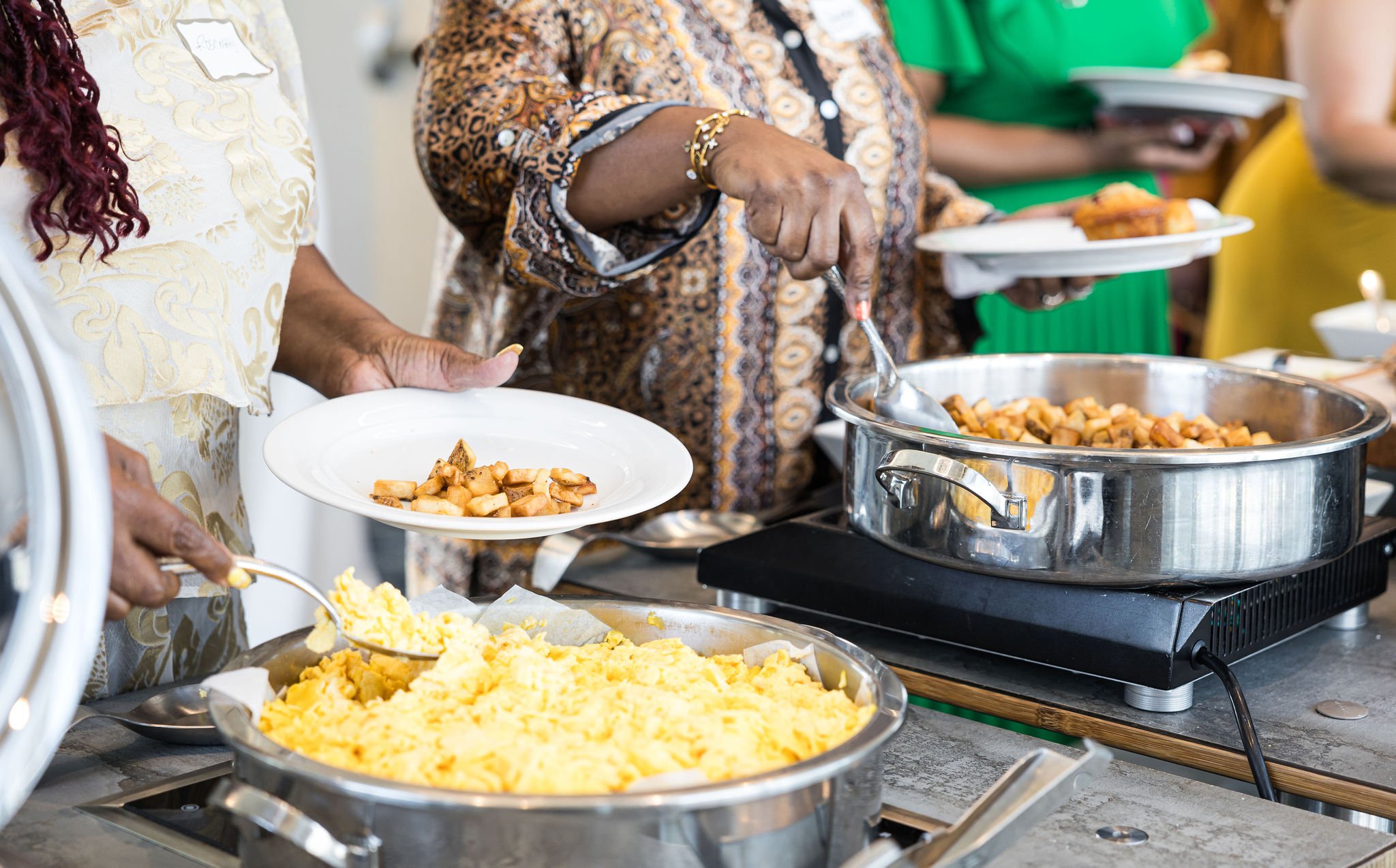 People serving scrambled eggs and potatoes from buffet containers.