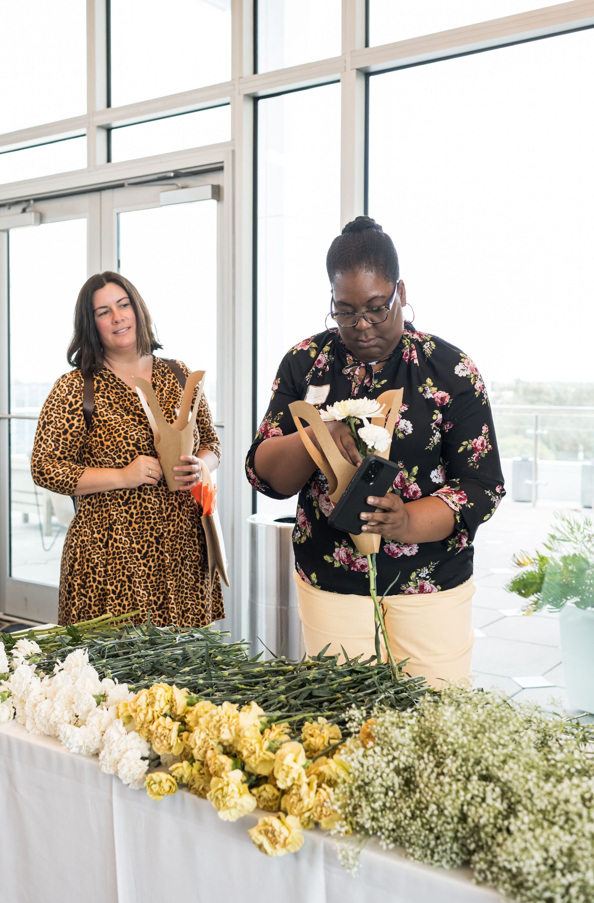Two women in floral dresses making bouquets at a table with white and yellow flowers in a bright room.