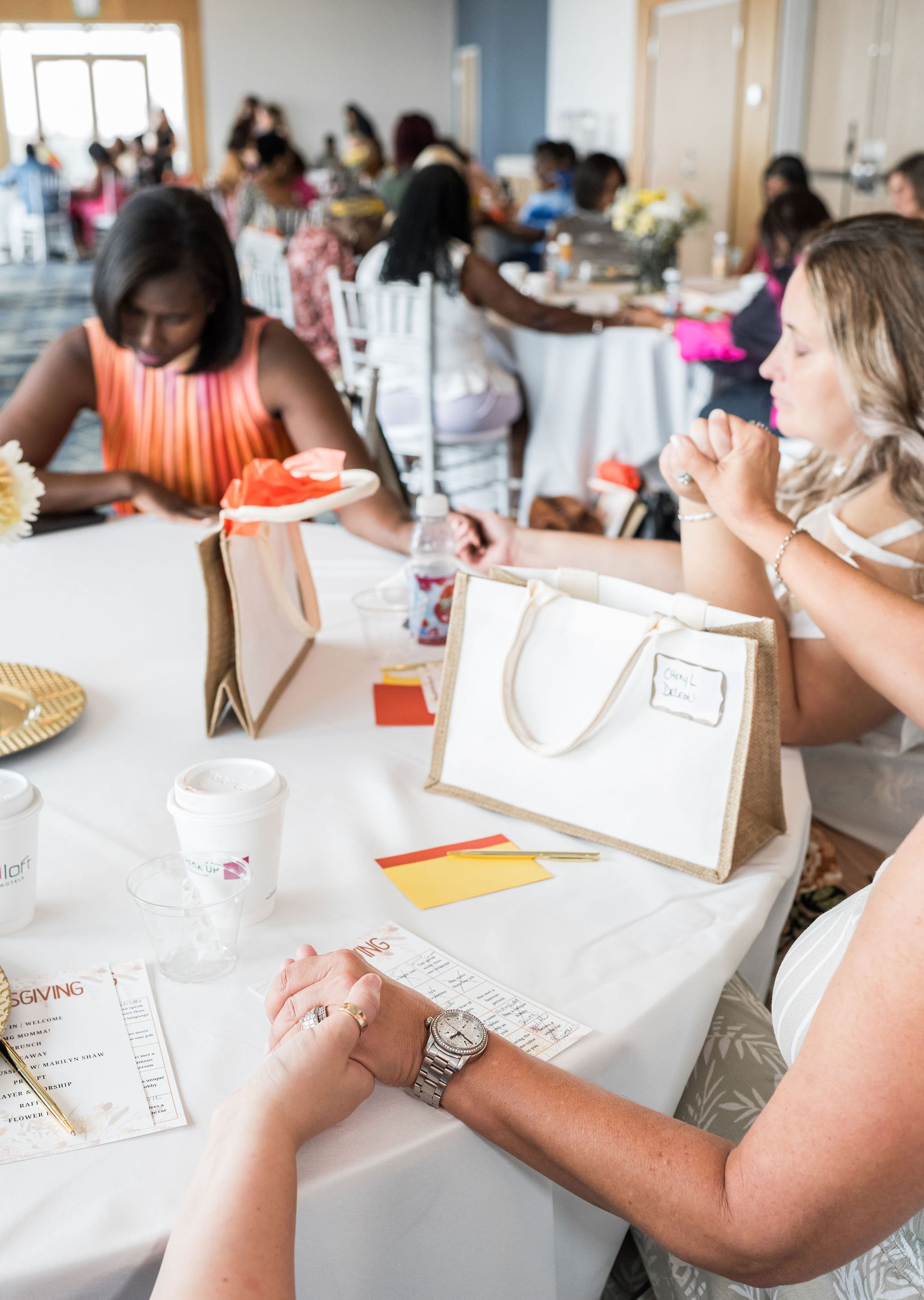 Group of people sitting at round tables with gift bags, paper cups, and holding hands in a meeting or event setting.