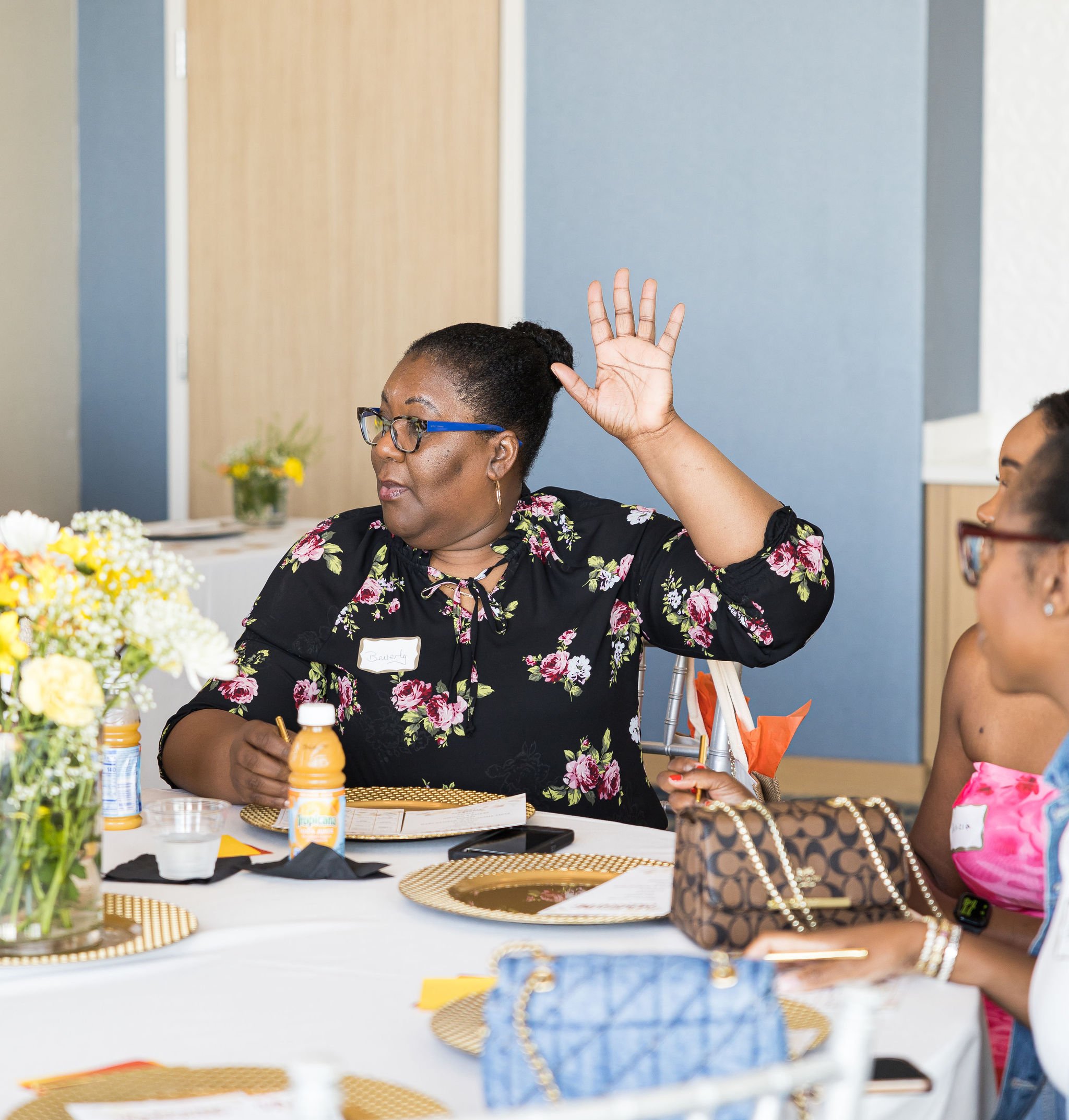 A woman in a floral blouse sitting at a table raising her hand, surrounded by others and decorative tableware. The table has flowers, plates, and beverages on it. The setting appears to be a social or professional gathering.