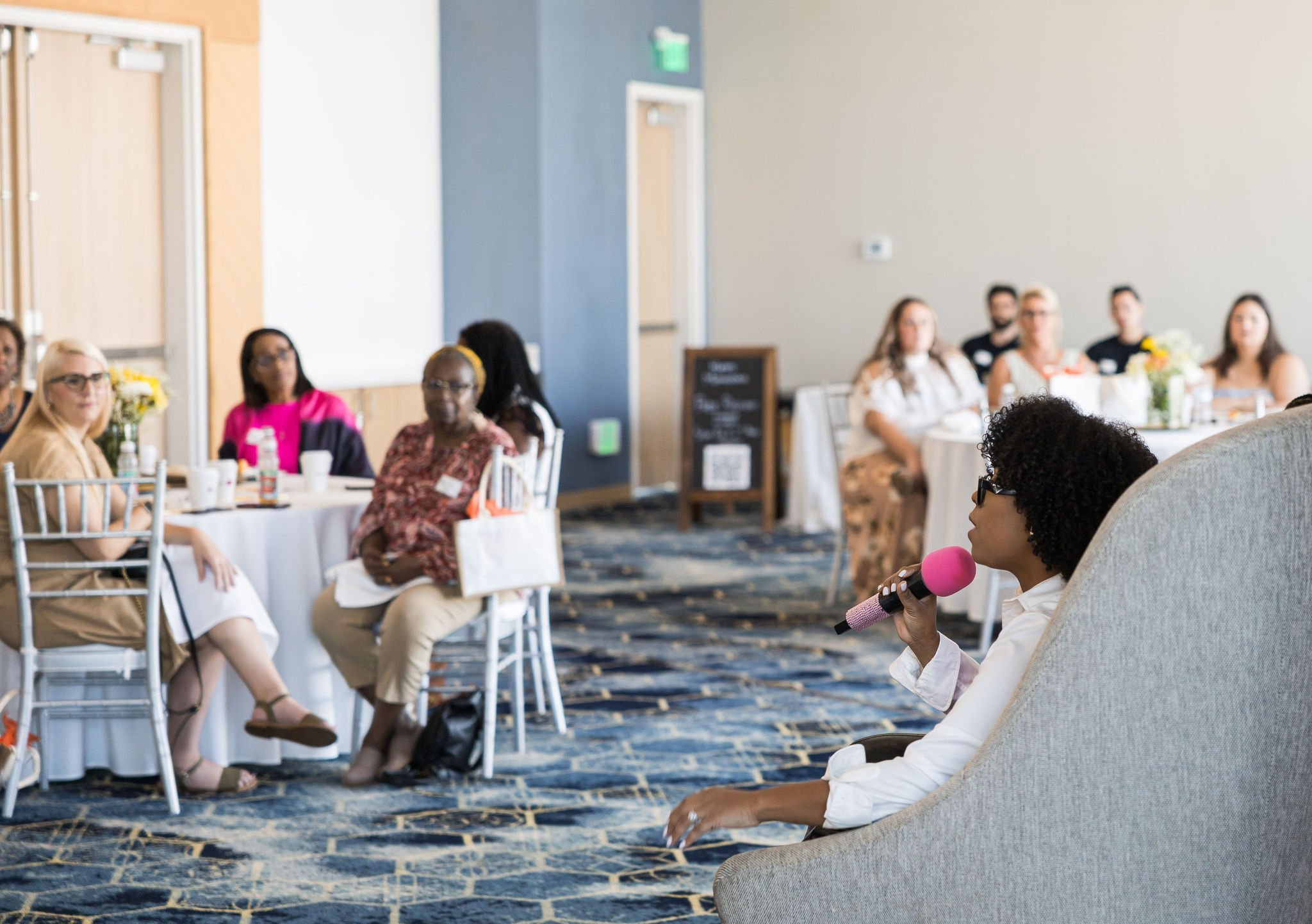 People sitting at tables in a conference room, one person holding a pink microphone, engaging in discussion.