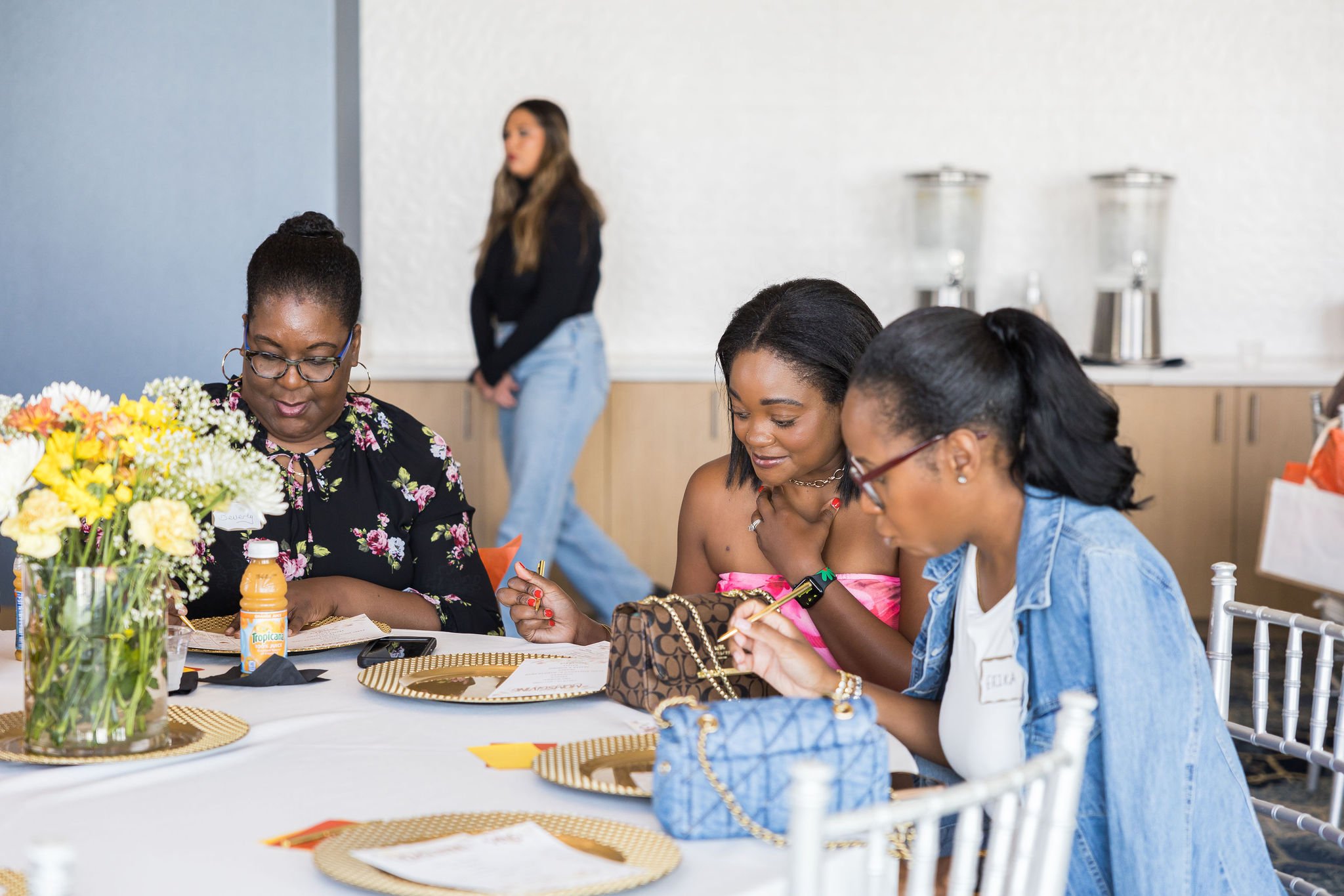 Three women sitting at a table with floral decorations, each focused on writing, with drinks and handbags nearby. A woman is standing in the background.