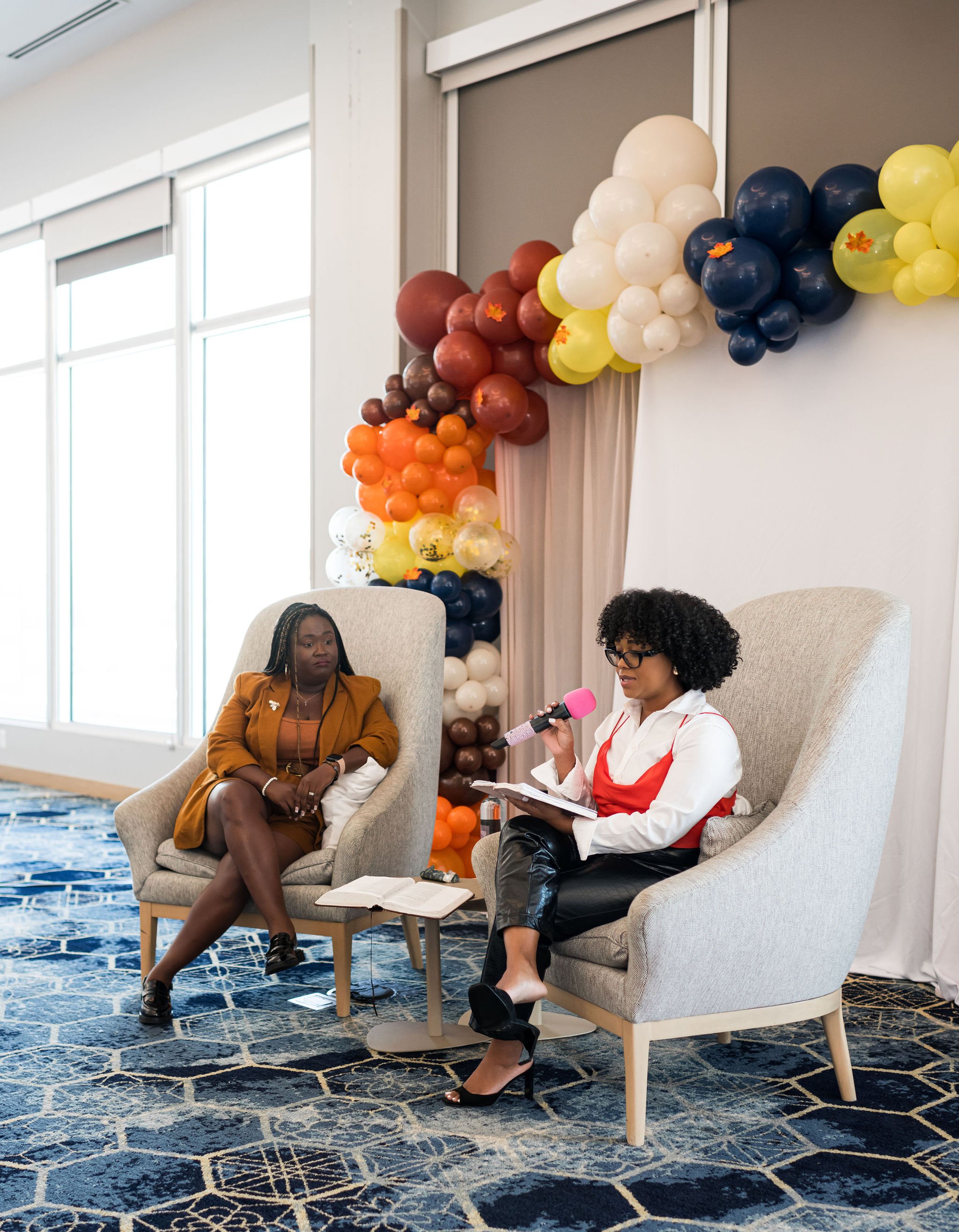 Two women seated in armchairs at an indoor event, with one holding a pink microphone and reading from a notebook. They are in front of a colorful balloon arch. The setting has large windows and a blue patterned carpet.