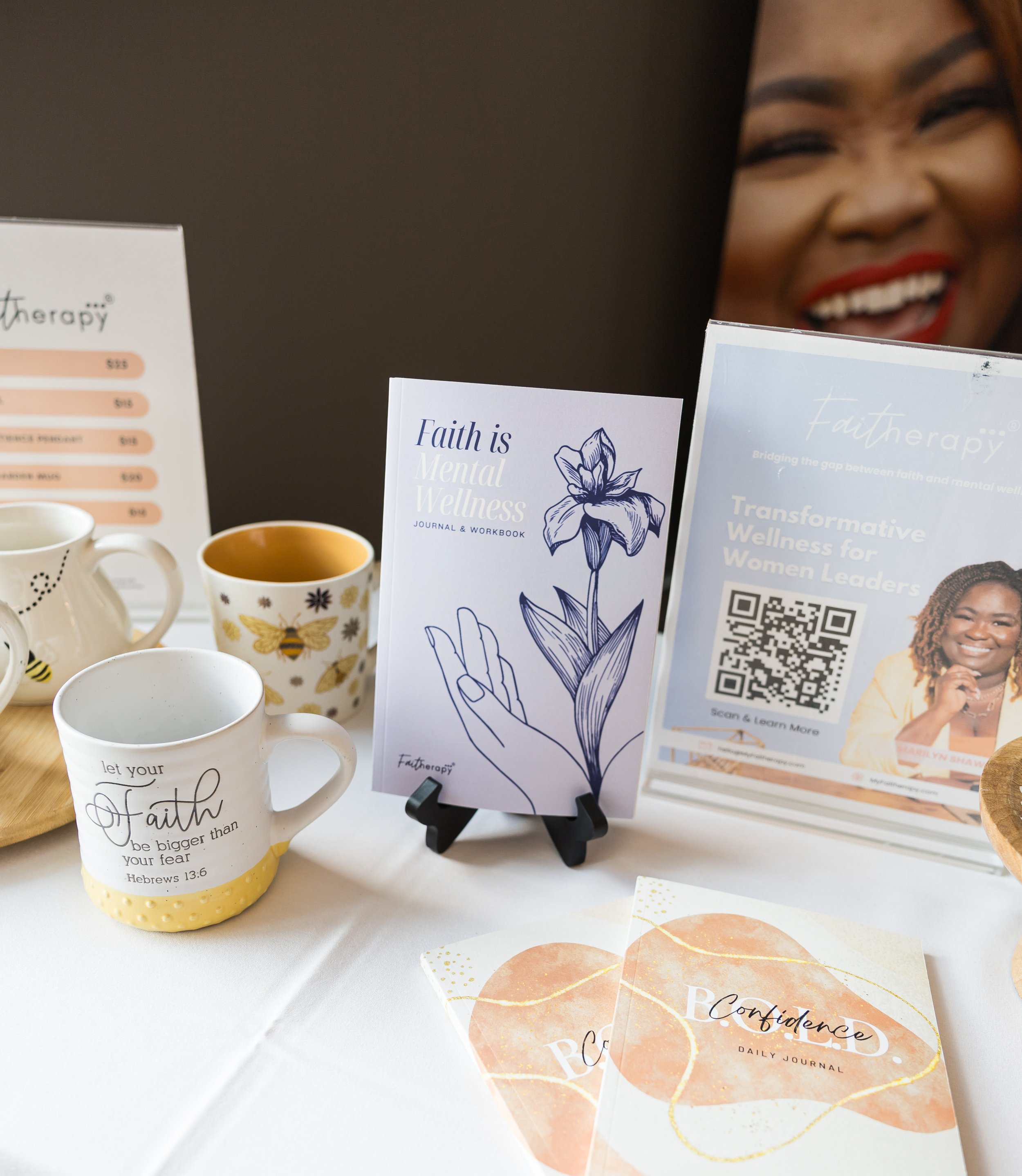 Display table with a 'Faith is Mental Wellness' journal, a mug with an inspirational quote, a 'Confidence Daily Journal,' and promotional materials. A smiling person's photo on a poster is in the background.