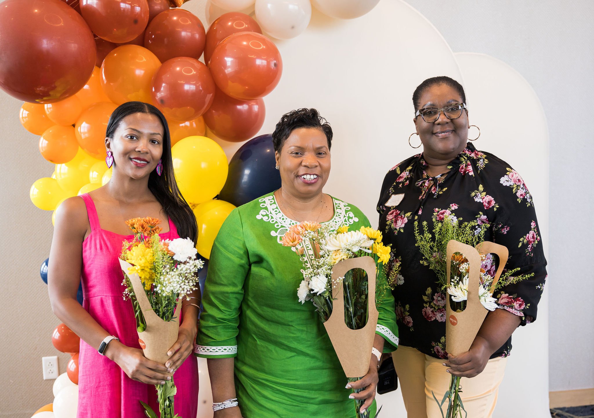 Three women holding bouquets standing in front of a colorful balloon arch with shades of orange, yellow, and white.