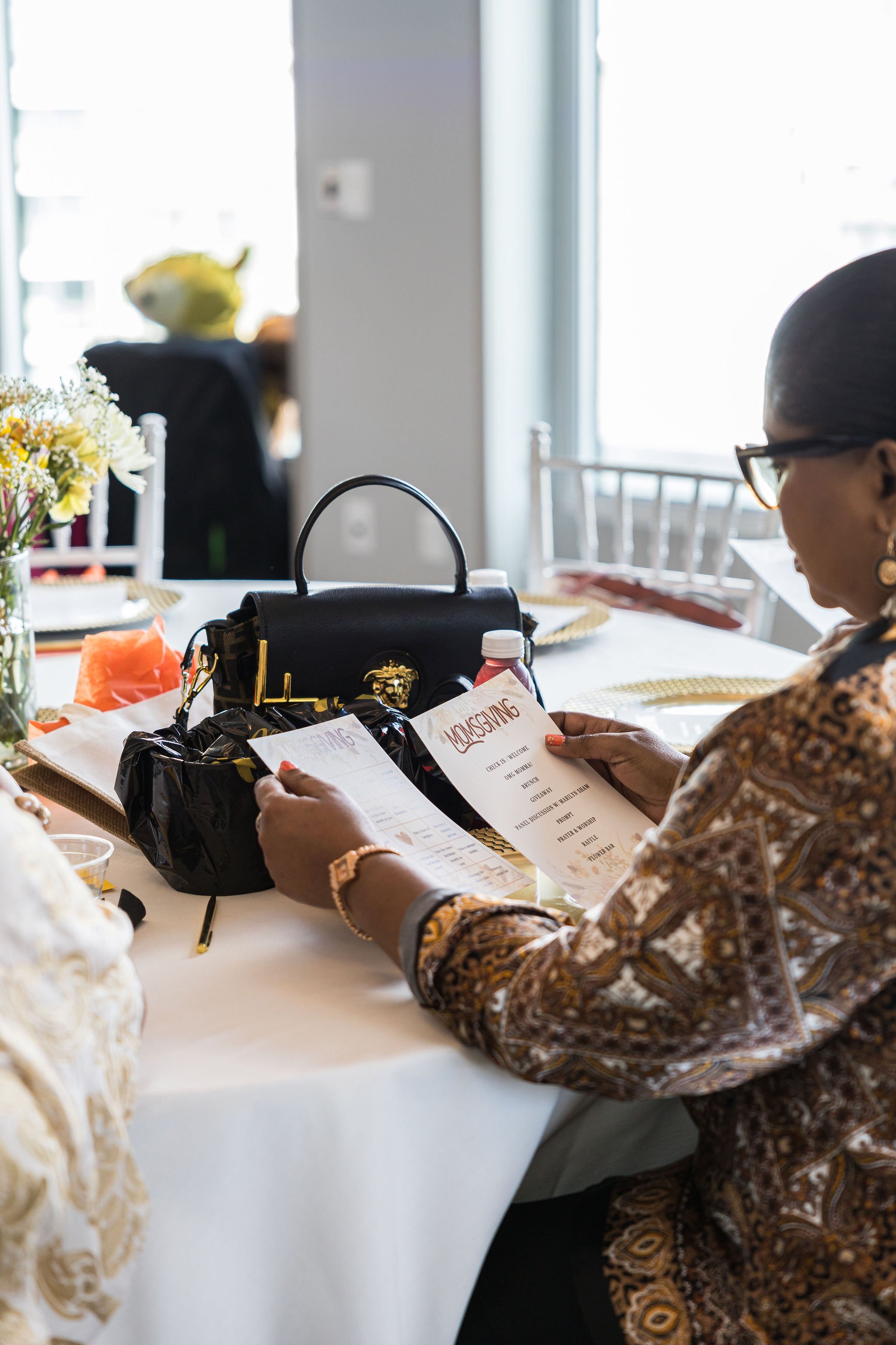 Person holding a menu labeled "Friendsgiving" at a table with a handbag, flowers, and plates.