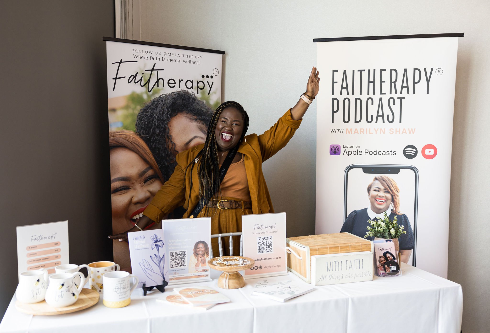 Woman smiling and raising hands beside Faitherapy podcast booth, featuring promotional materials and banners.