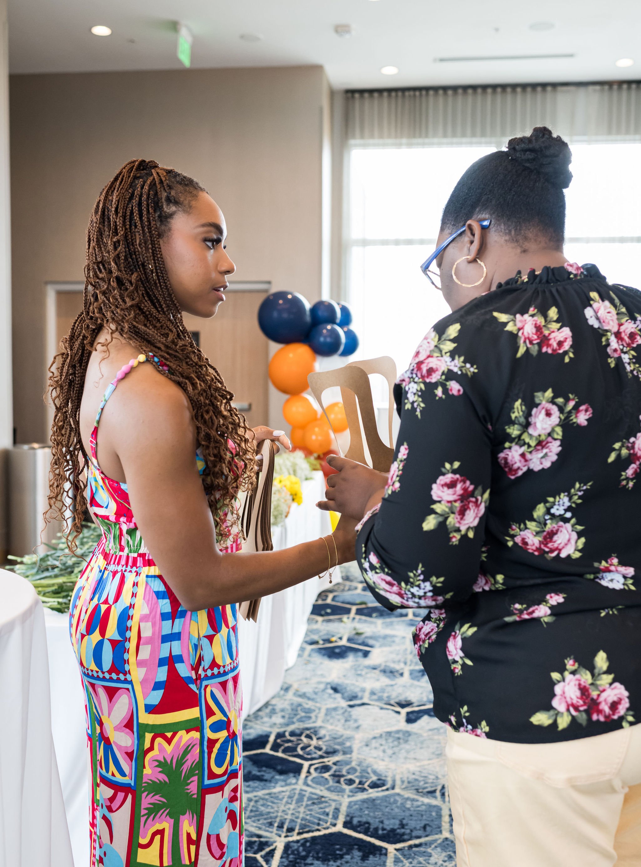 Two women in conversation, one wearing a colorful dress and the other in a floral top, standing in a decorated room with balloons.