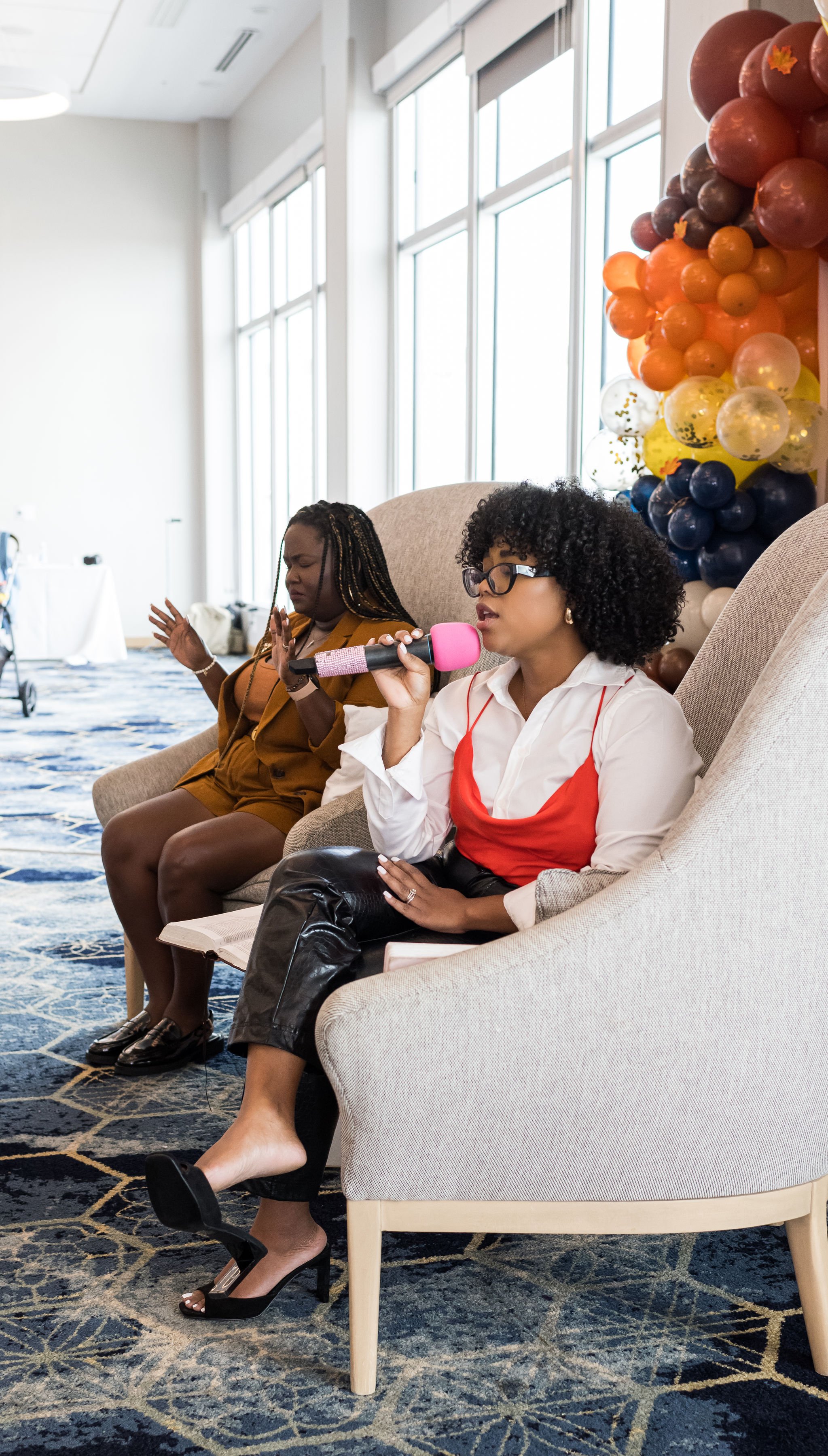 Two women sitting in chairs; one holding a pink microphone, wearing a white shirt and red top. The other is in a brown outfit with braided hair. Colorful balloon arrangement in the background.