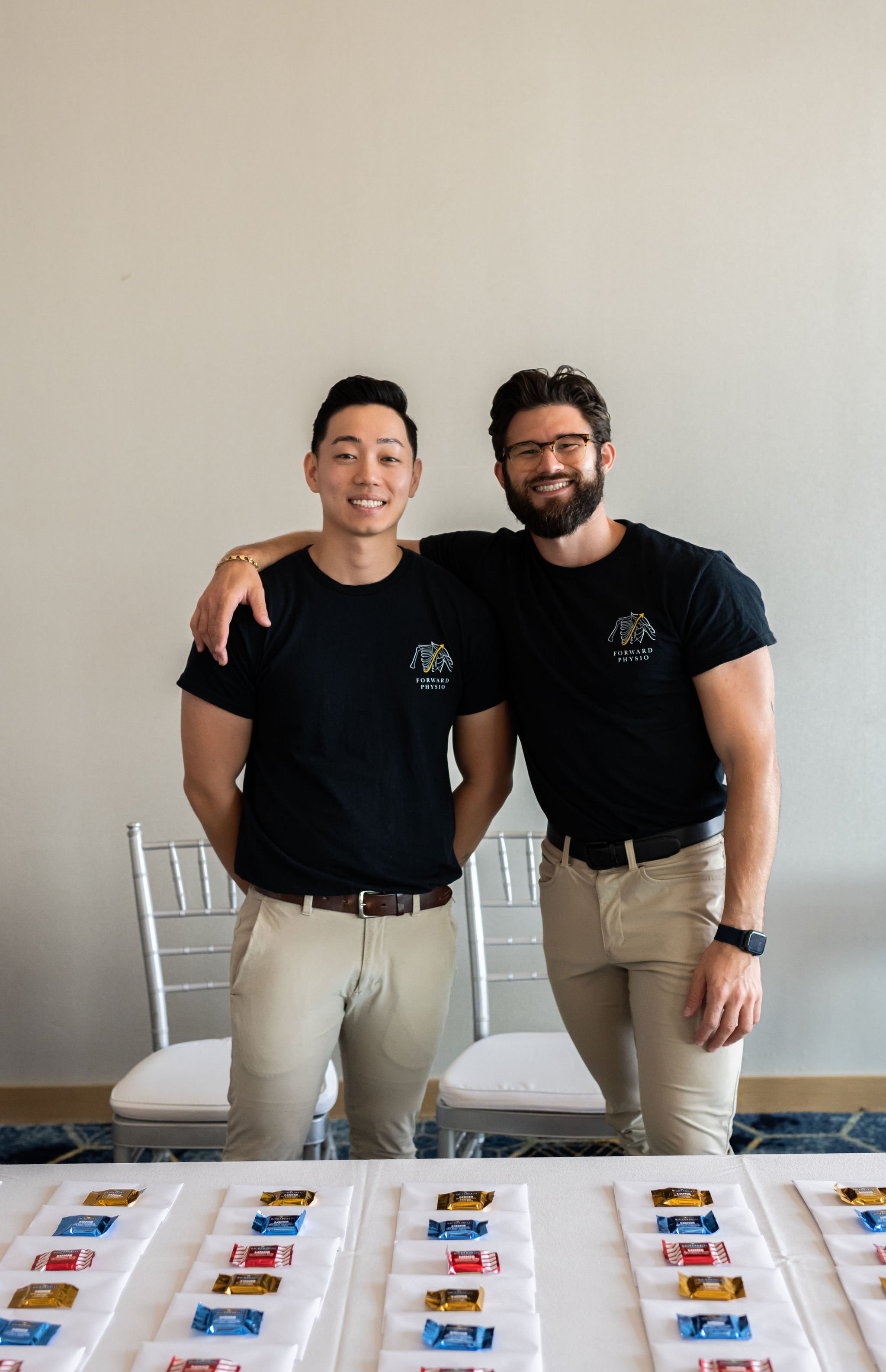 Two men smiling and posing together, both wearing black t-shirts with "Forward Physio" logos, standing behind a table with arranged small candy packets on napkins. There are empty chairs in the background.