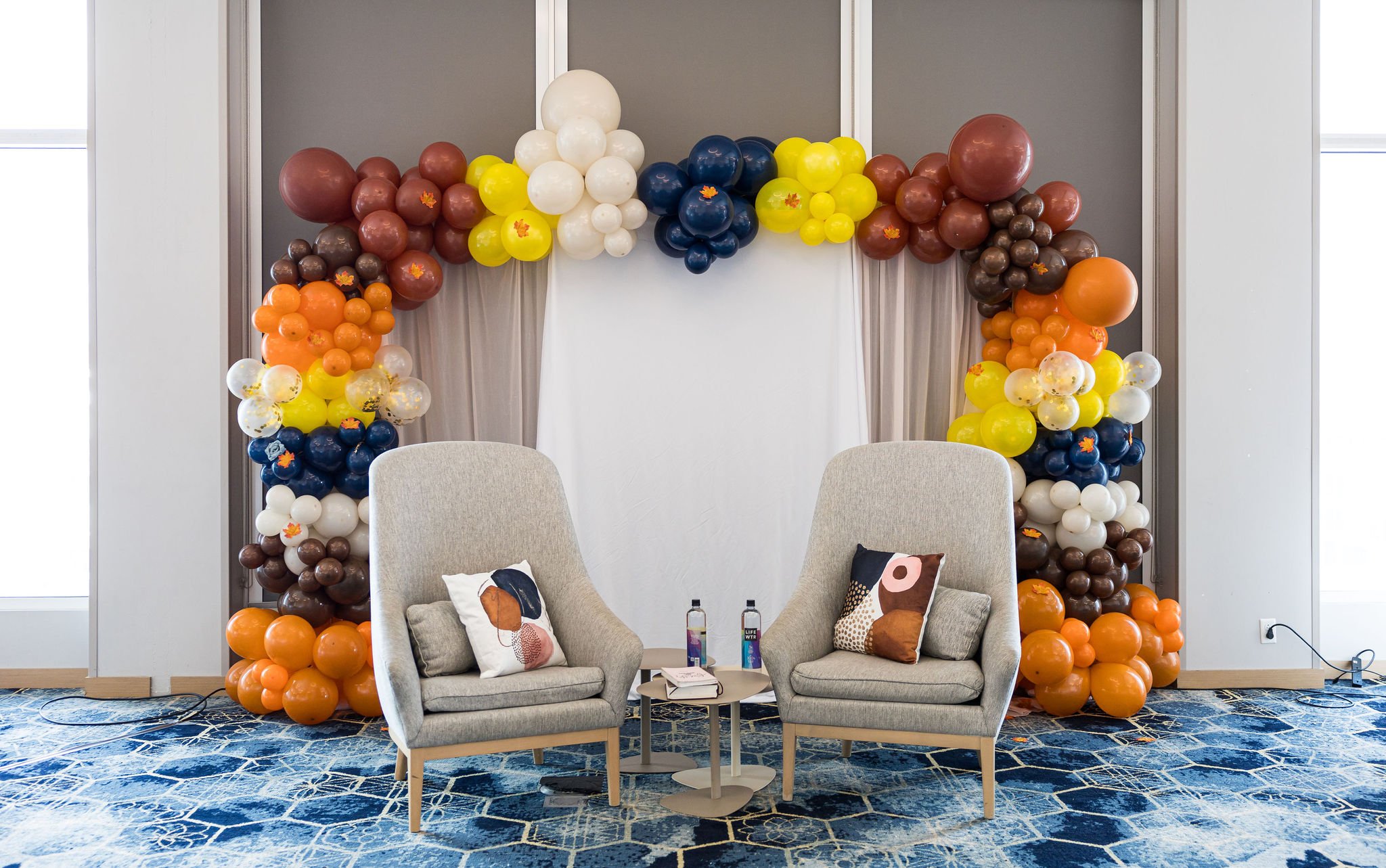 Decorative balloon arch with two beige chairs and a small table, featuring colorful balloons in orange, yellow, and navy blue, with fall-themed accents. The chairs have decorative pillows and bottled water on the table, set on a patterned blue carpet