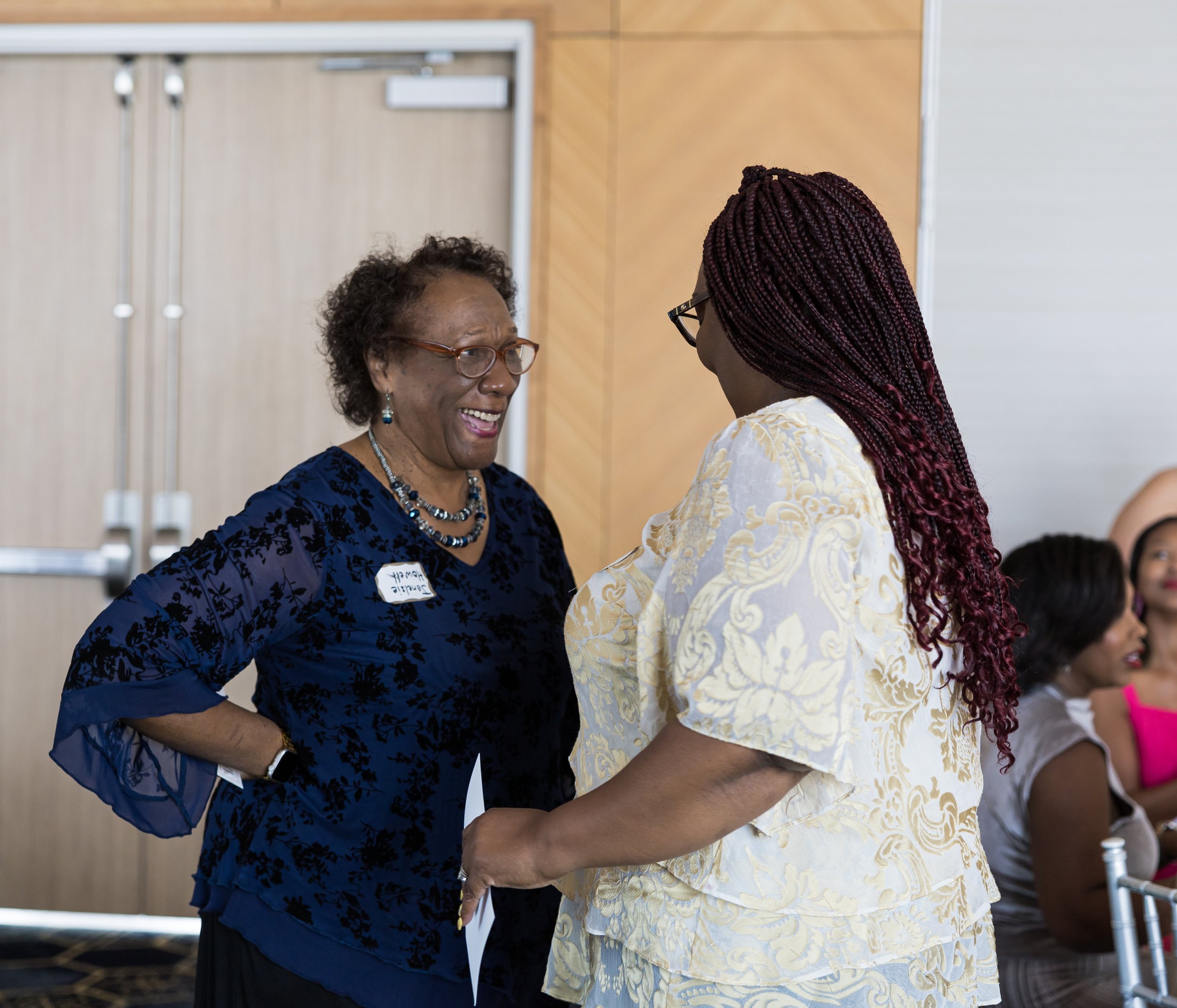 Two women talking and smiling at an indoor event, with a group of seated people in the background.