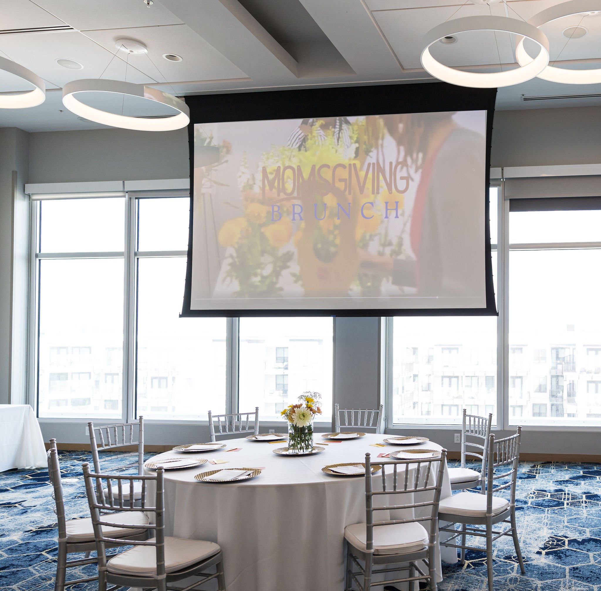 Conference room set up for a brunch event with round tables, white tablecloths, chairs, and a video screen displaying "Momsgiving Brunch."