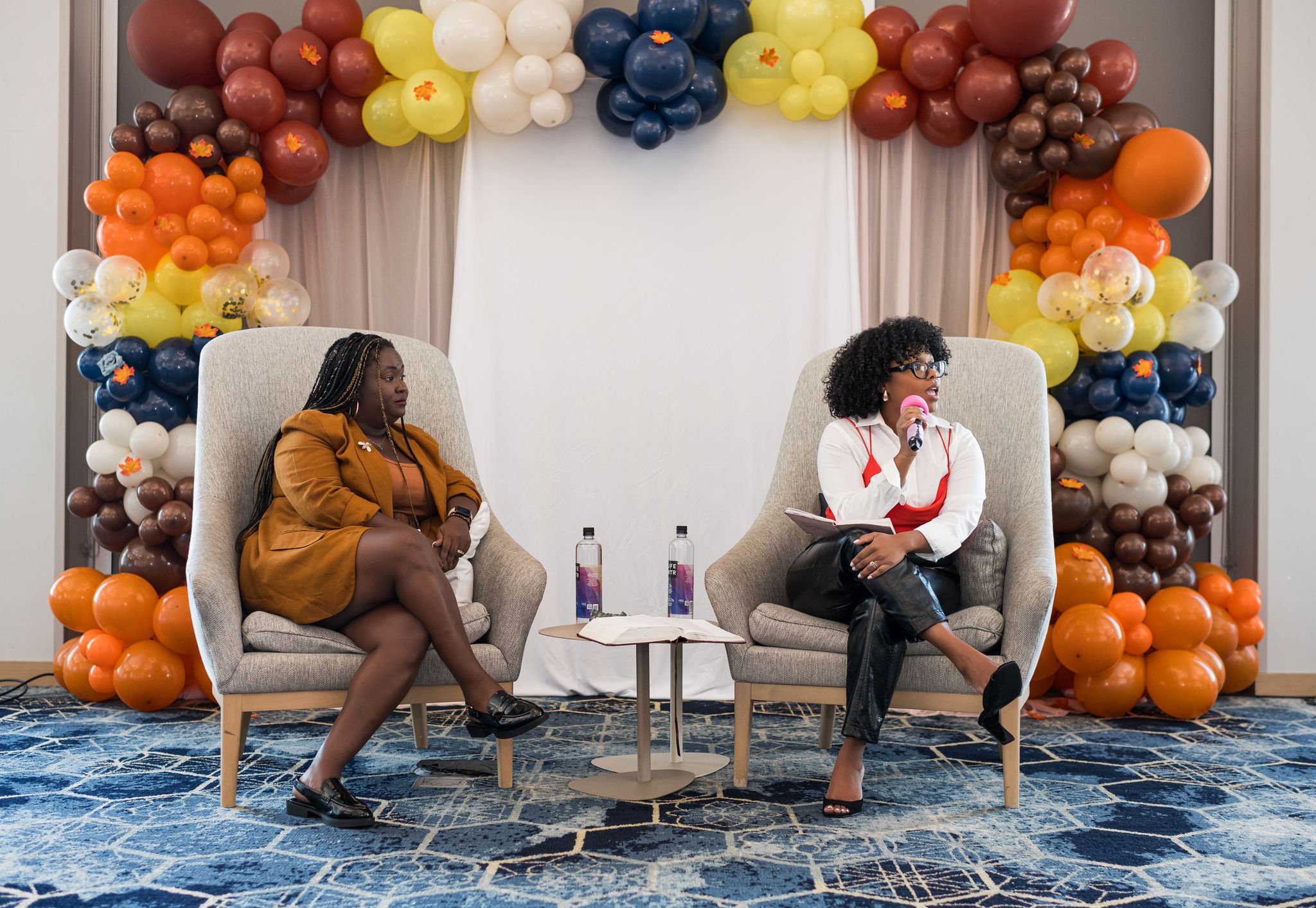 Two women seated on armchairs, engaged in a discussion on stage. They are surrounded by colorful balloon arrangements in fall colors like orange, yellow, brown, and white. One holds a microphone while the other is listening attentively. A small table