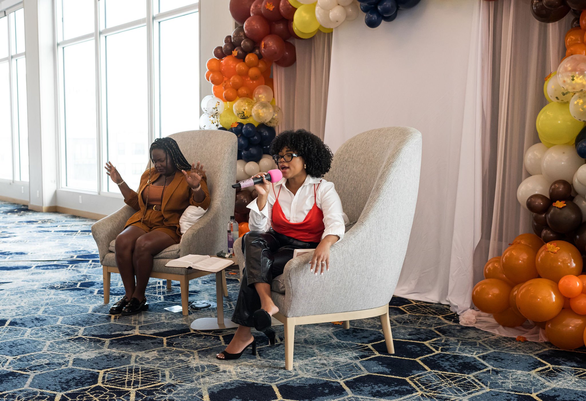 Two women seated in armchairs during an event with balloon decorations in the background. One woman holds a microphone speaking, while the other has her hands raised. Both are on a patterned carpet.