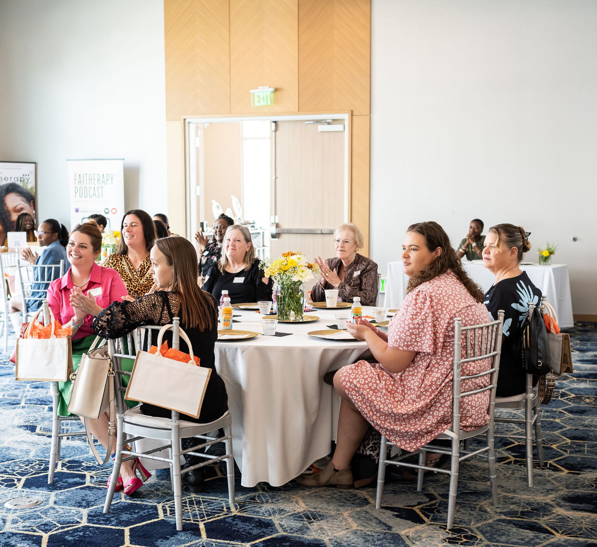 Group of women sitting around a round table at an indoor event, with gift bags and flower centerpiece on the table.