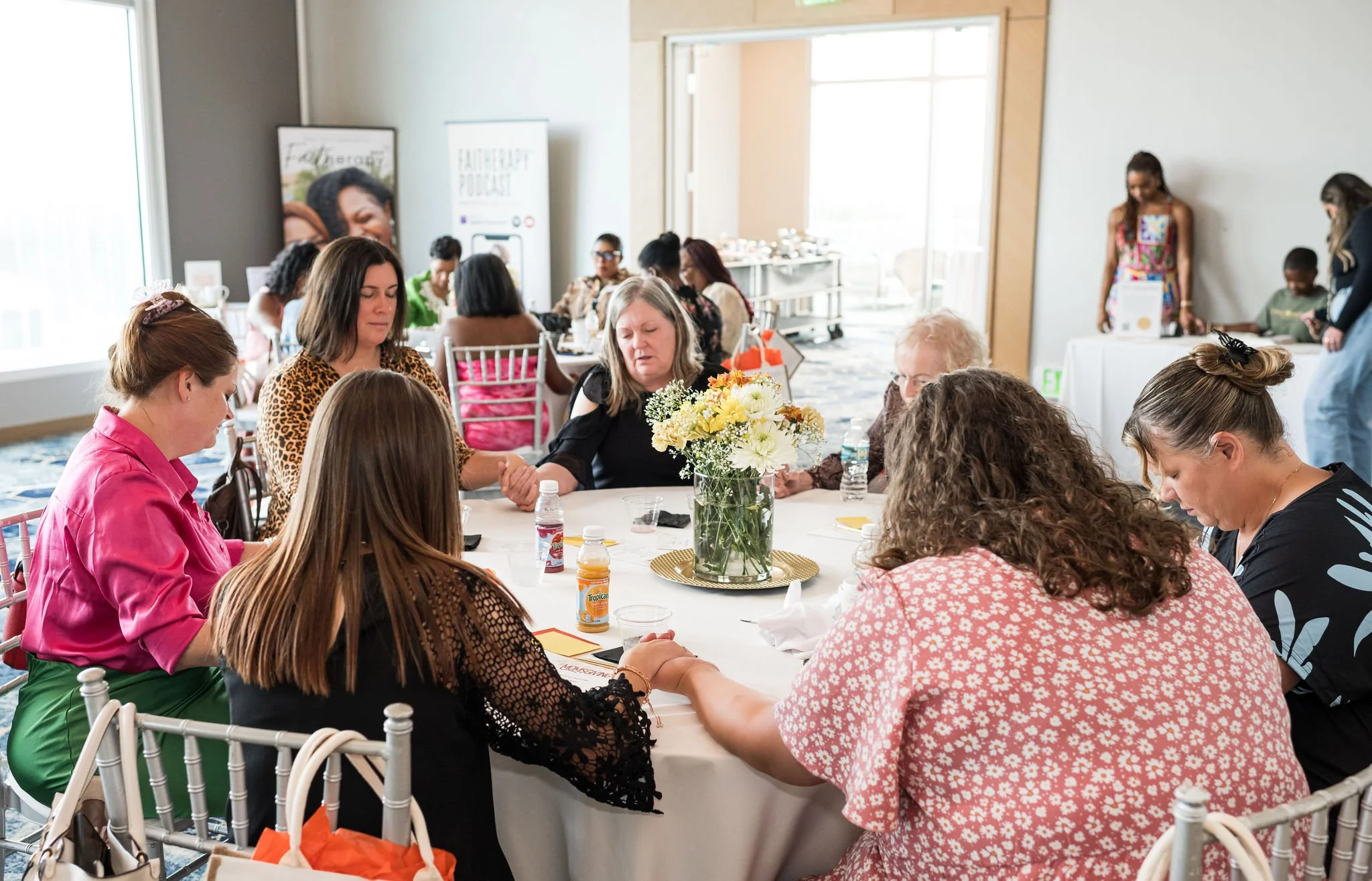 A group of women sitting around a table, holding hands in prayer. The table is decorated with flowers and juice bottles. A banner in the background promotes a faith-themed podcast.