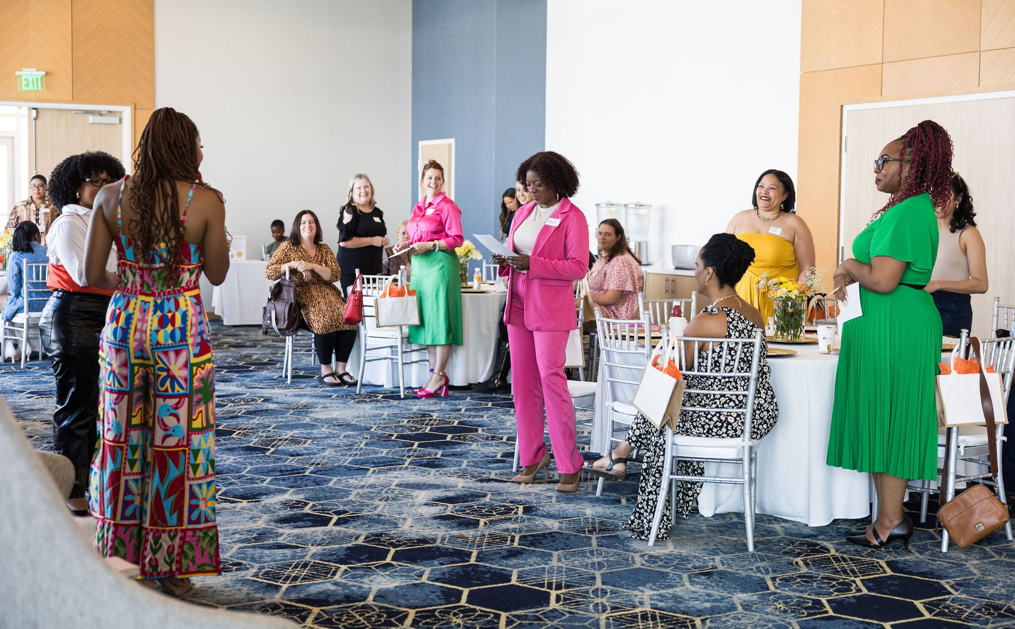 A group of diverse women standing and sitting around tables in a brightly lit room, dressed in vibrant outfits, engaged in a social or professional gathering.