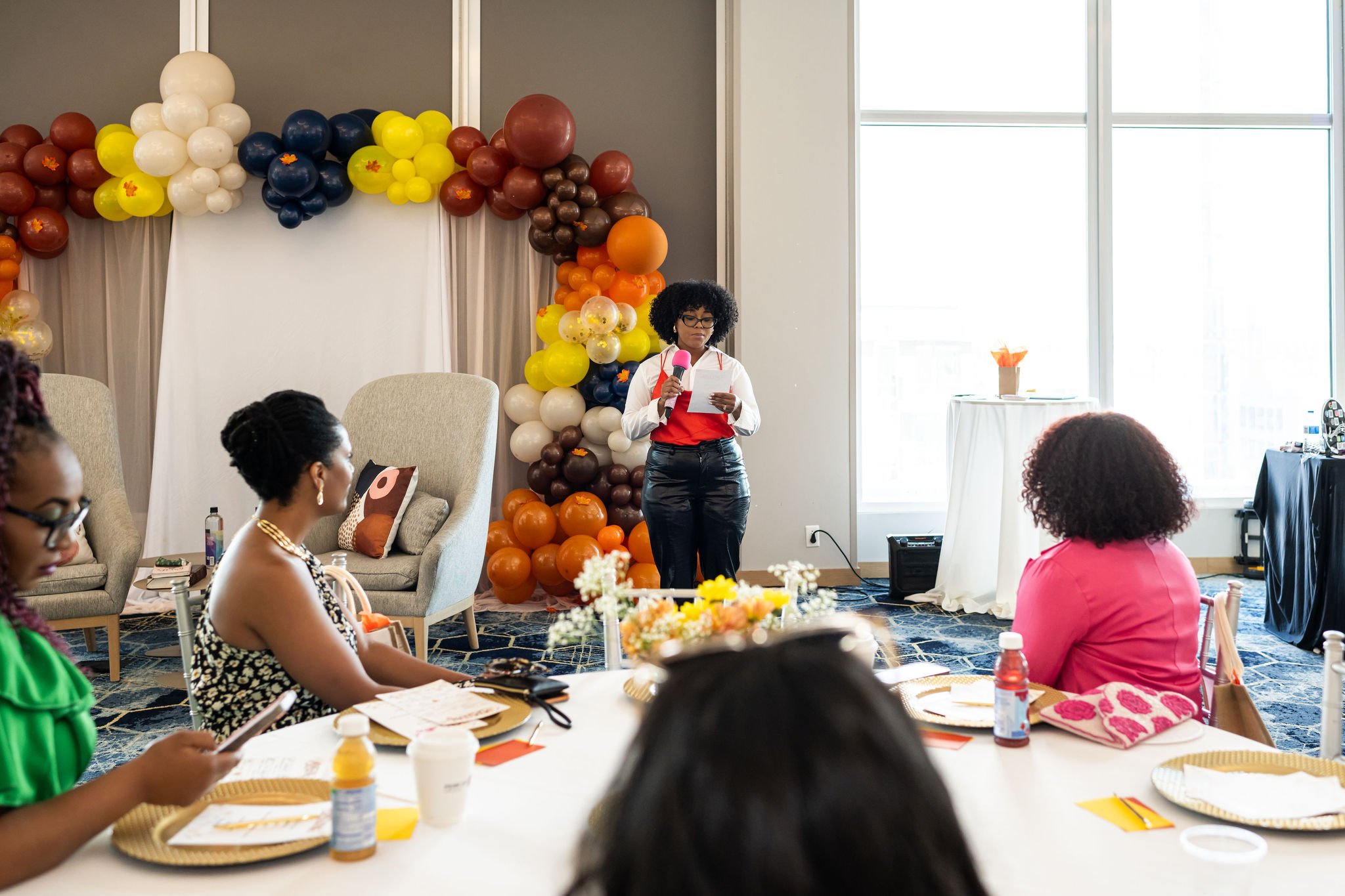 A woman speaking with a pink microphone at an indoor event, surrounded by colorful balloon decorations and seated attendees at a table with food and drinks.