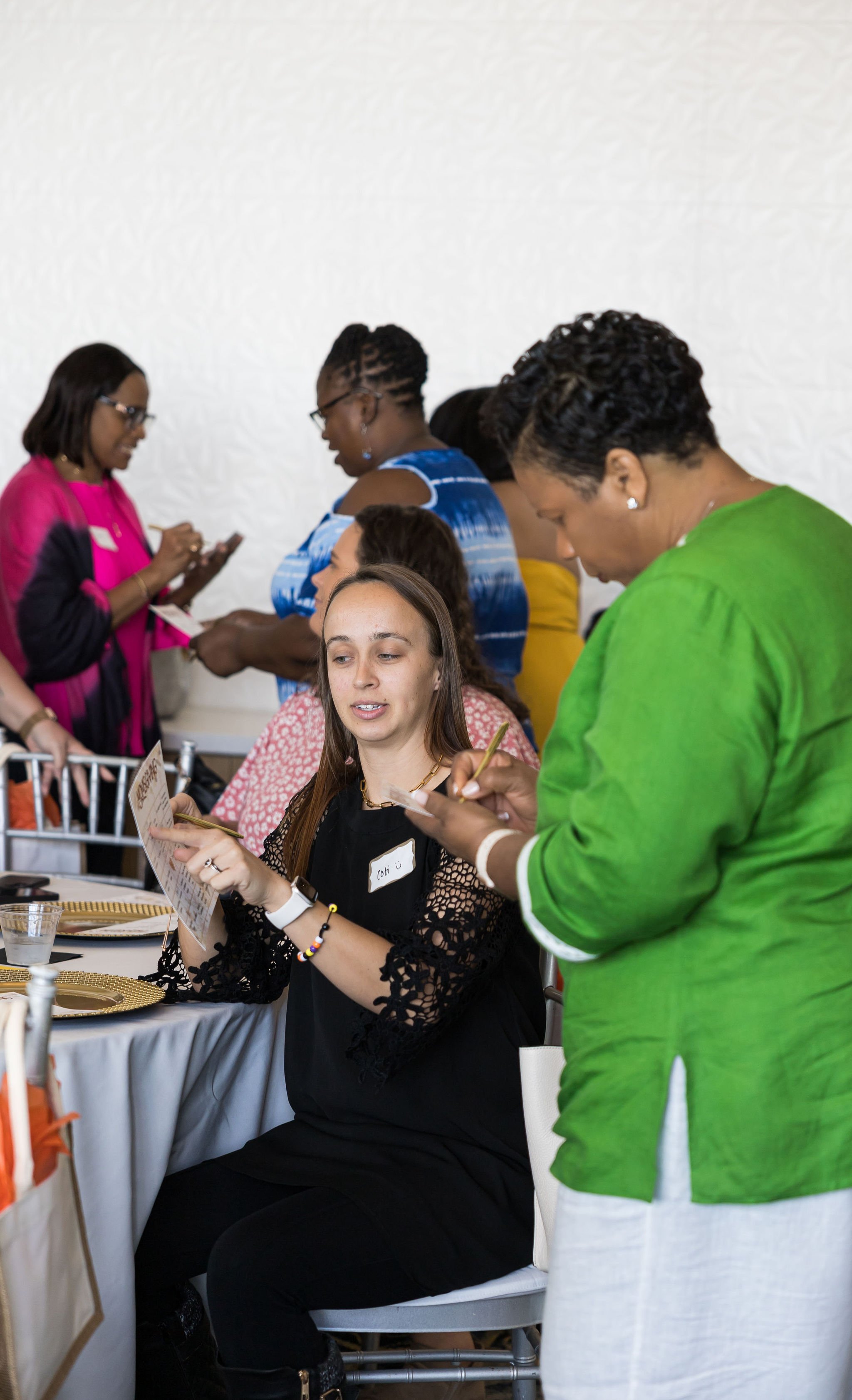 A group of people at an event, with one person seated at a table holding a paper, surrounded by others standing and engaging in conversation, all dressed in colorful clothing.