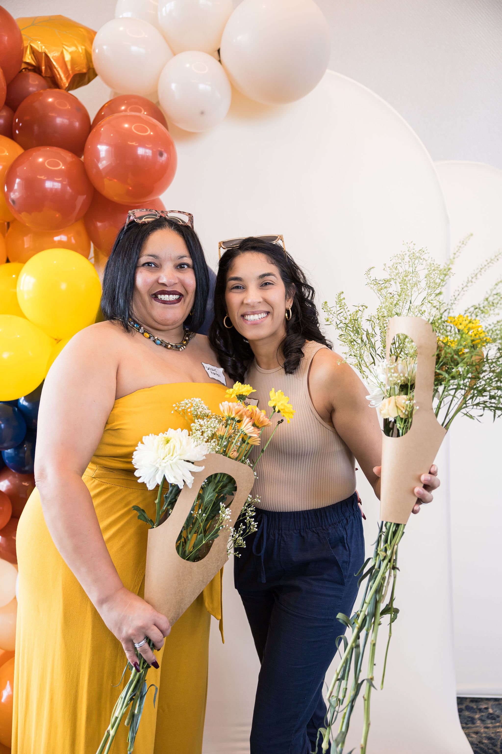 Two people smiling, holding bouquets wrapped in brown paper, standing in front of a colorful balloon backdrop.