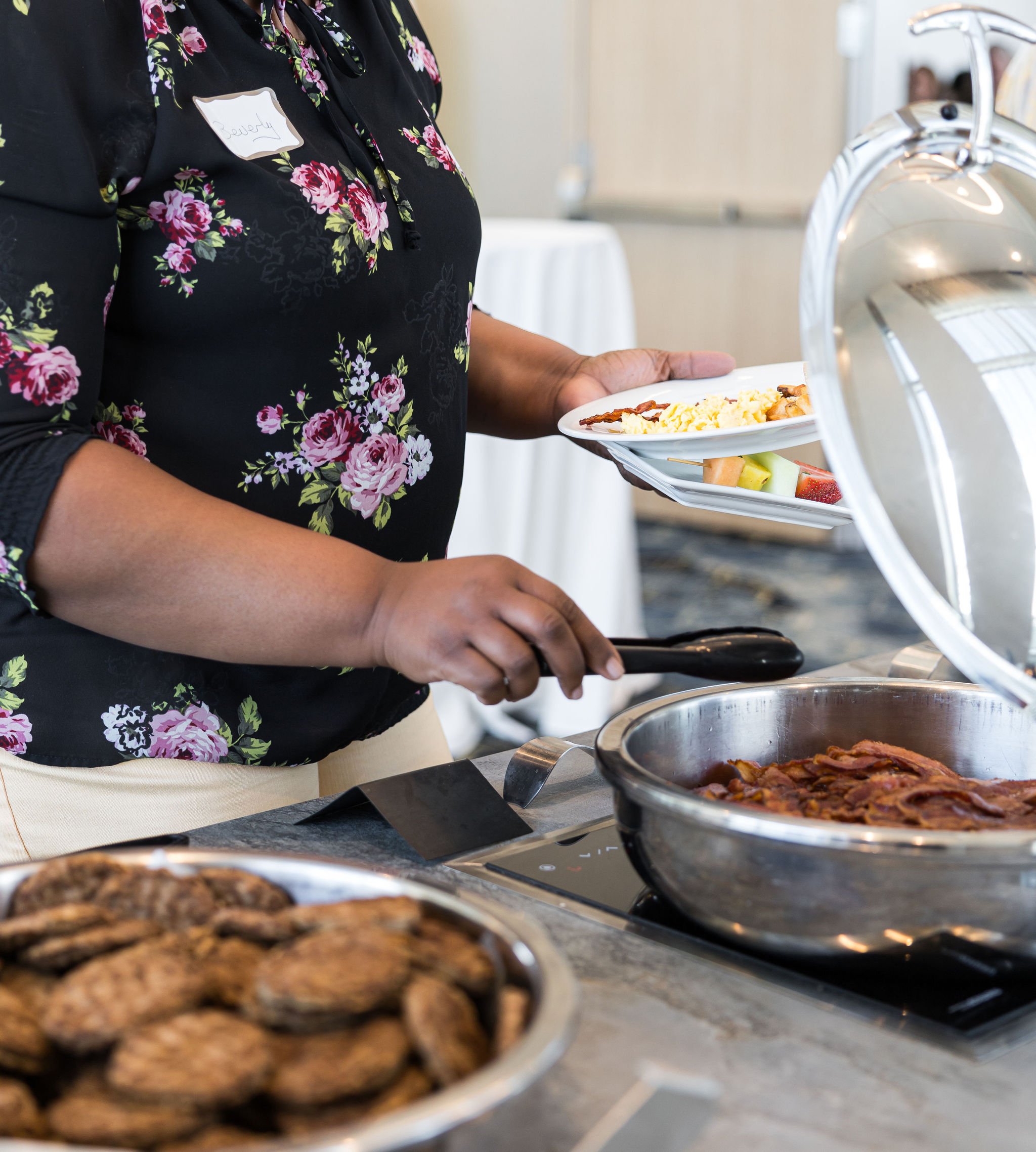 Person serving food from a buffet station with a stainless steel chafing dish containing bacon, next to a plate of cookies.