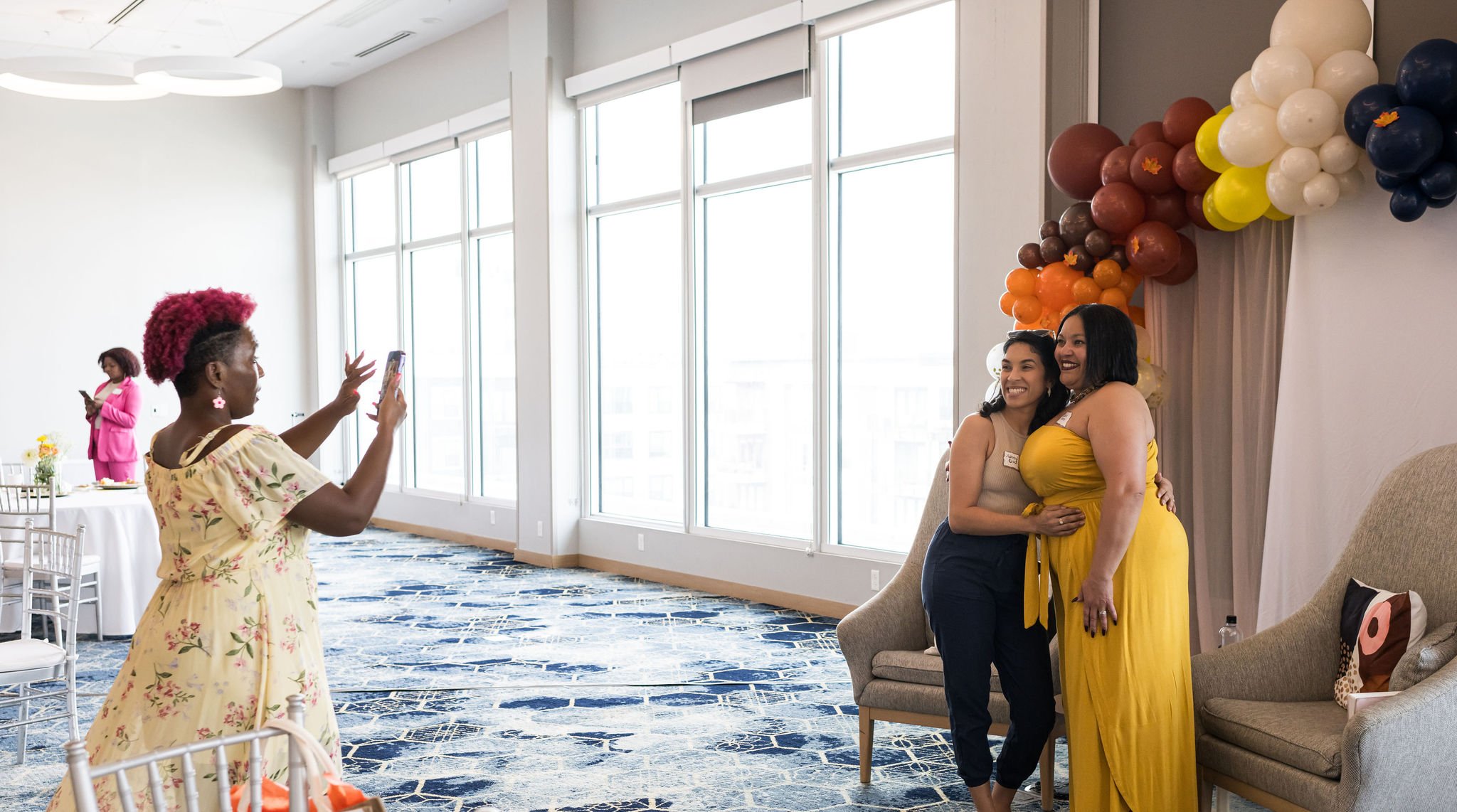 A group of people at a gathering with two women posing for a photo in a well-lit room, decorated with colorful balloons, and another person taking the picture.