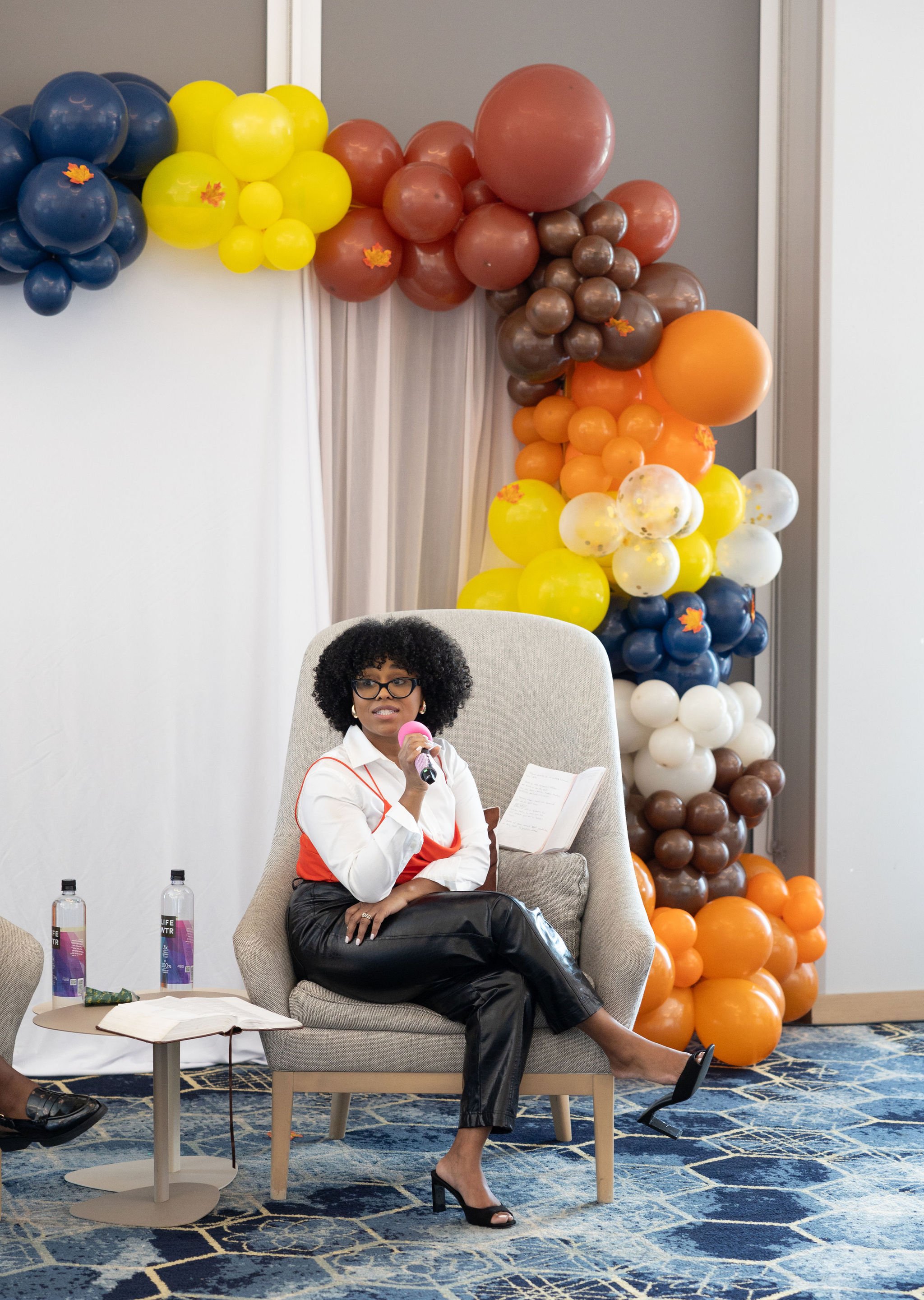 Person sitting in a chair holding a microphone, with a colorful balloon arch in the background. Two water bottles are on a small table nearby. The person is wearing glasses, a white shirt, and black pants.