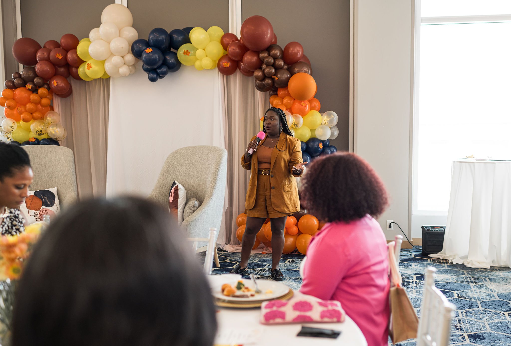 A person in a brown outfit speaking with a microphone in front of a seated audience. Behind them is a colorful balloon arch with shades of brown, red, yellow, and blue, flanking a white backdrop and armchair.