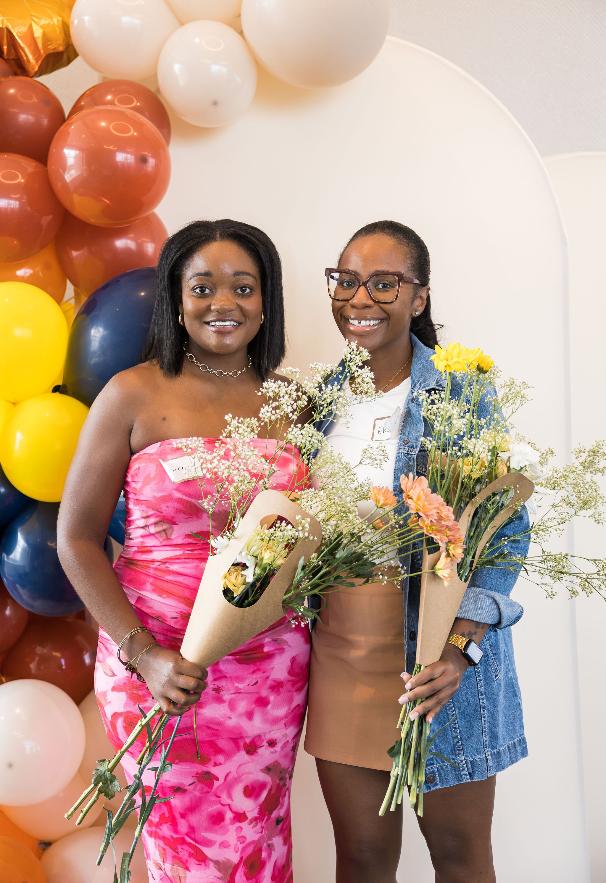 Two women holding bouquets of flowers, standing in front of a colorful balloon backdrop, smiling.