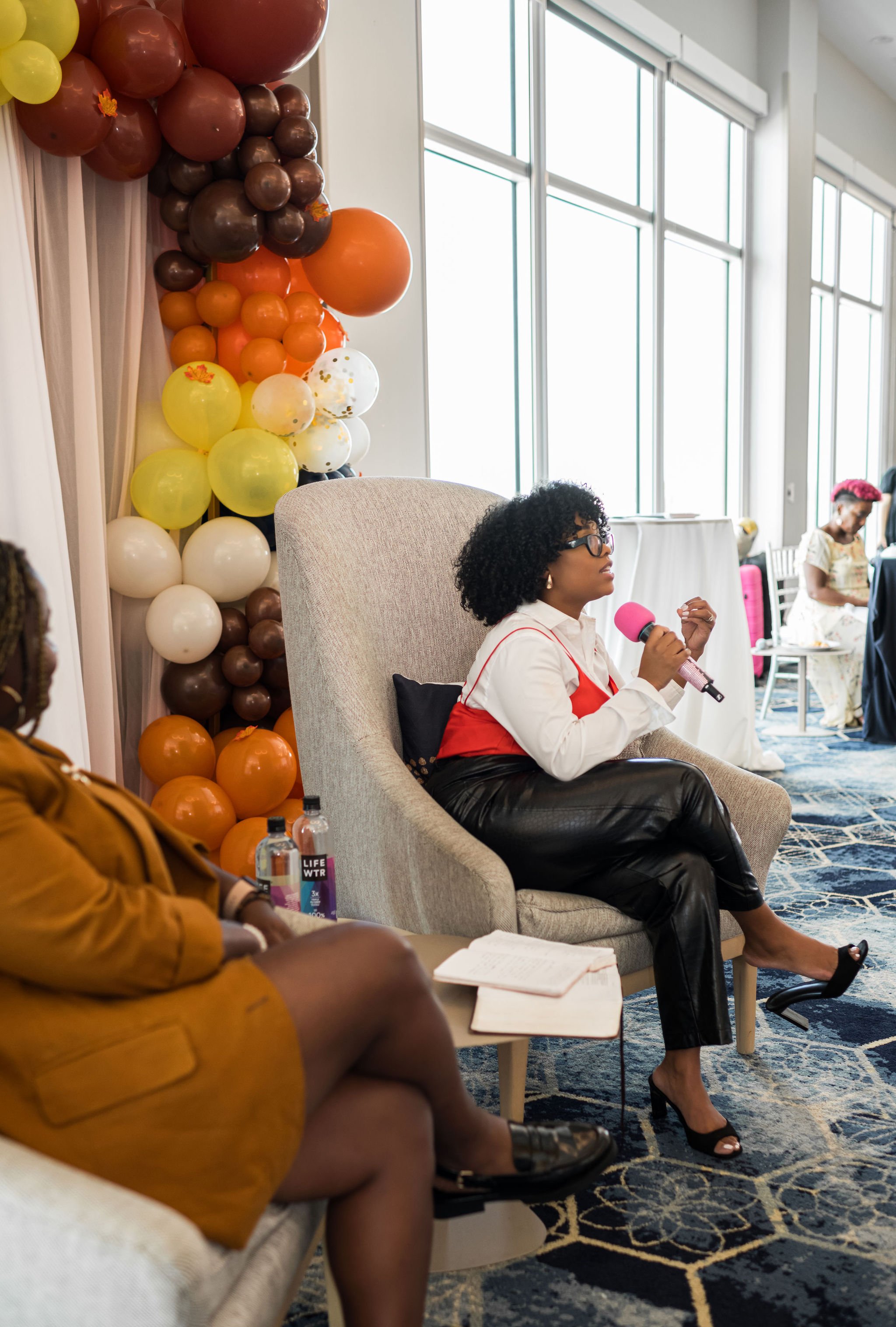 Two women seated in armchairs with a colorful balloon display in the background, one holding a pink microphone, engaged in conversation at an indoor event.