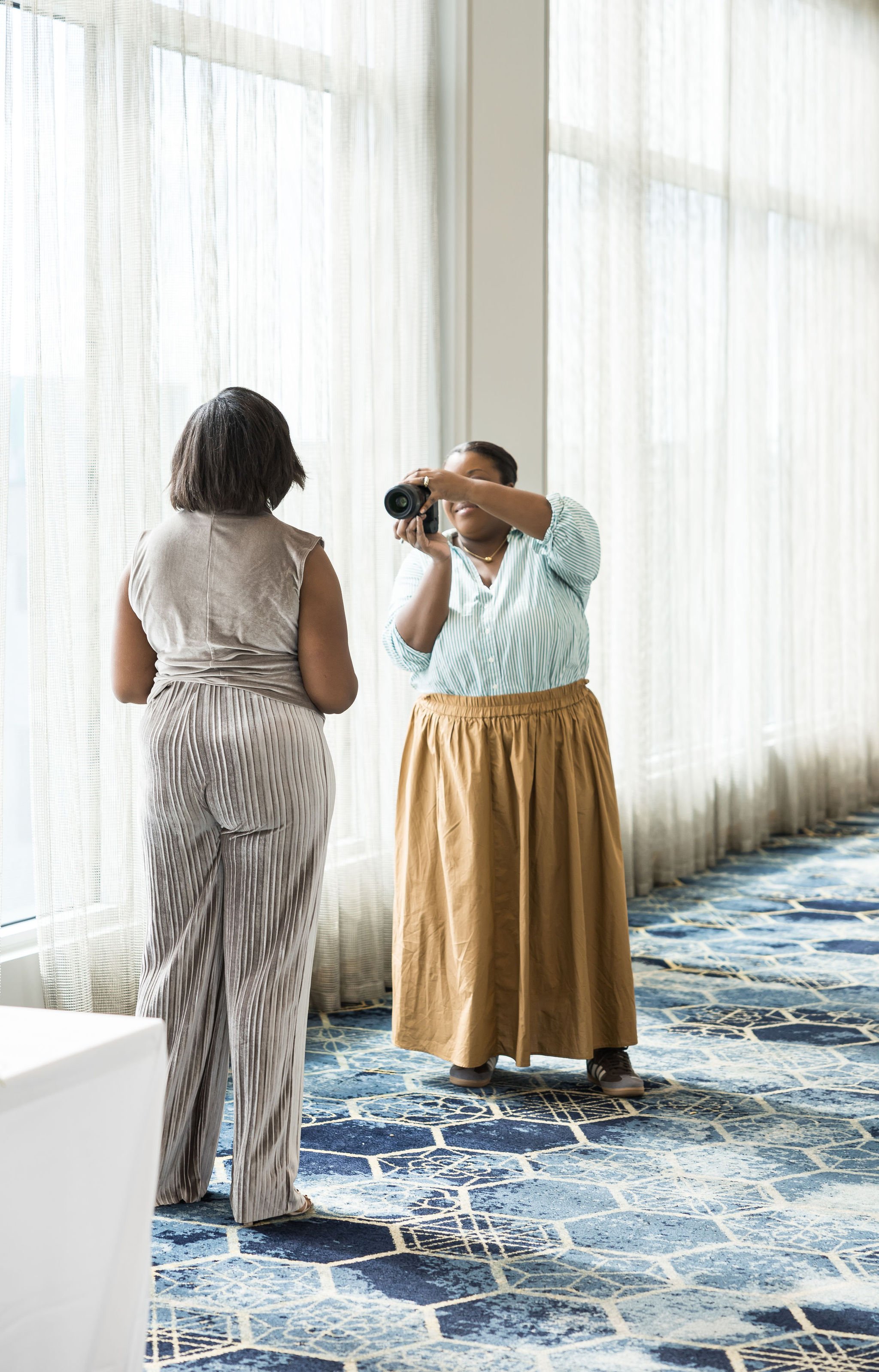 A photographer taking a picture of a person indoors, with both individuals standing on a patterned blue carpet. The photographer is wearing a light blue shirt and brown skirt, while the subject is wearing a sleeveless top and striped pants. The setti