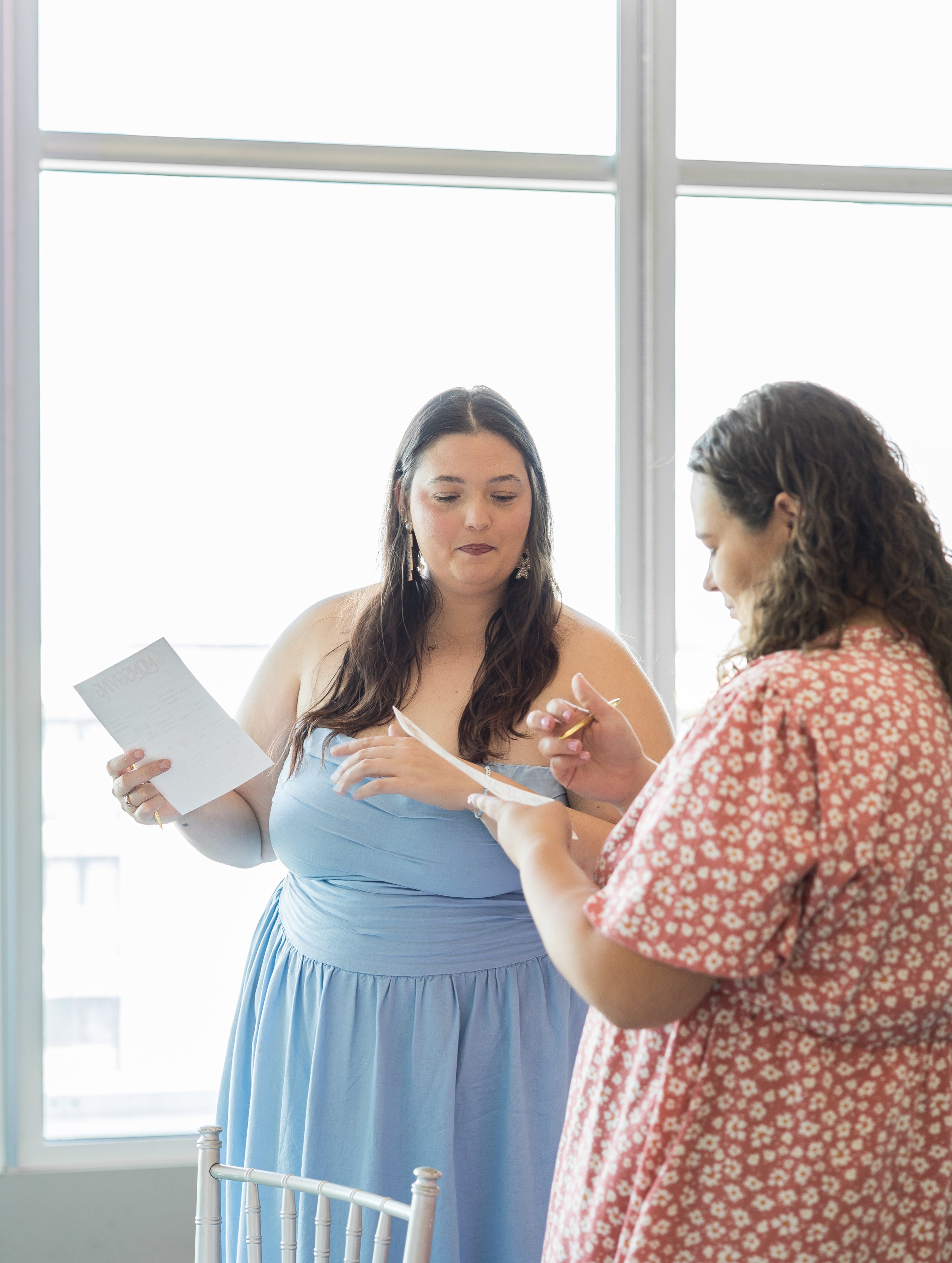 Two women standing indoors near a bright window, holding and looking at papers, engaged in a discussion.