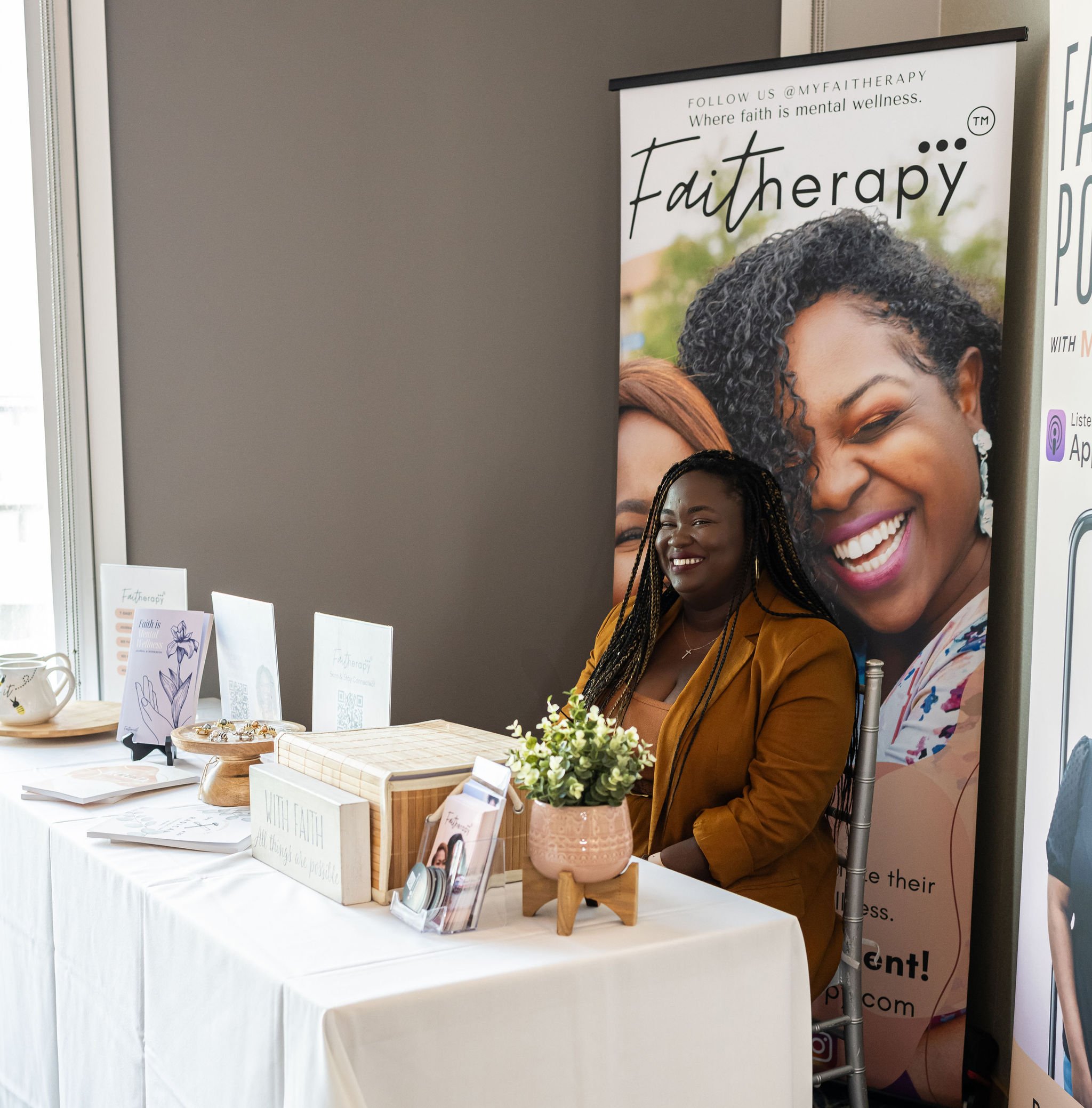 Woman sitting at a promotional booth for 'Faith Therapy' with brochures, a wooden box, and a pot of flowers on a table.