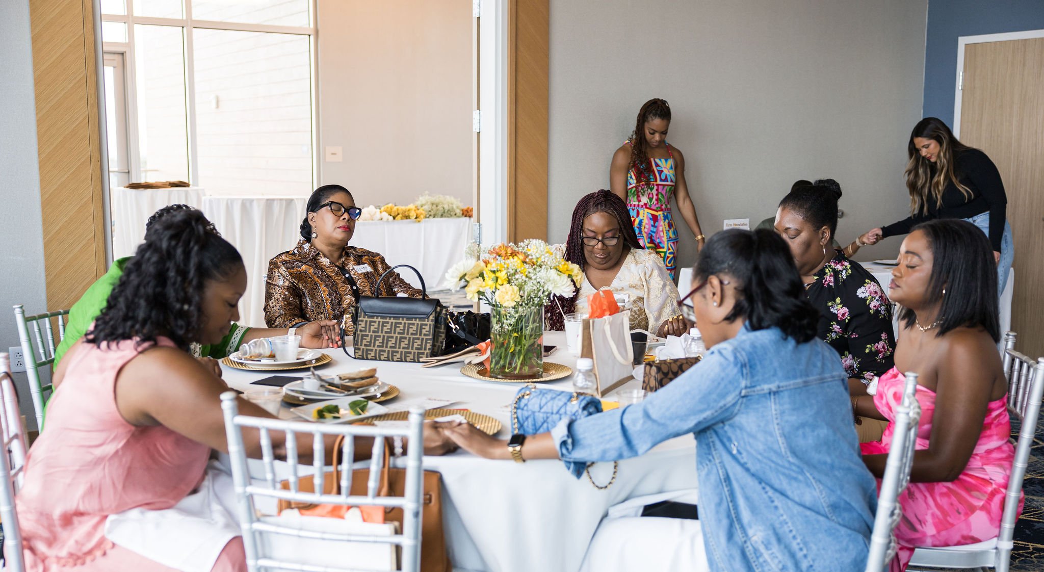 Women seated around a dining table, some holding hands, at a social gathering, with flowers at the center.