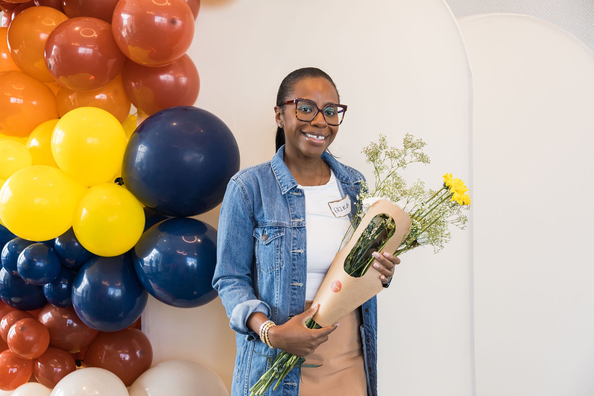 Woman wearing glasses and a denim jacket smiles while holding a bouquet of flowers, standing in front of colorful balloons.